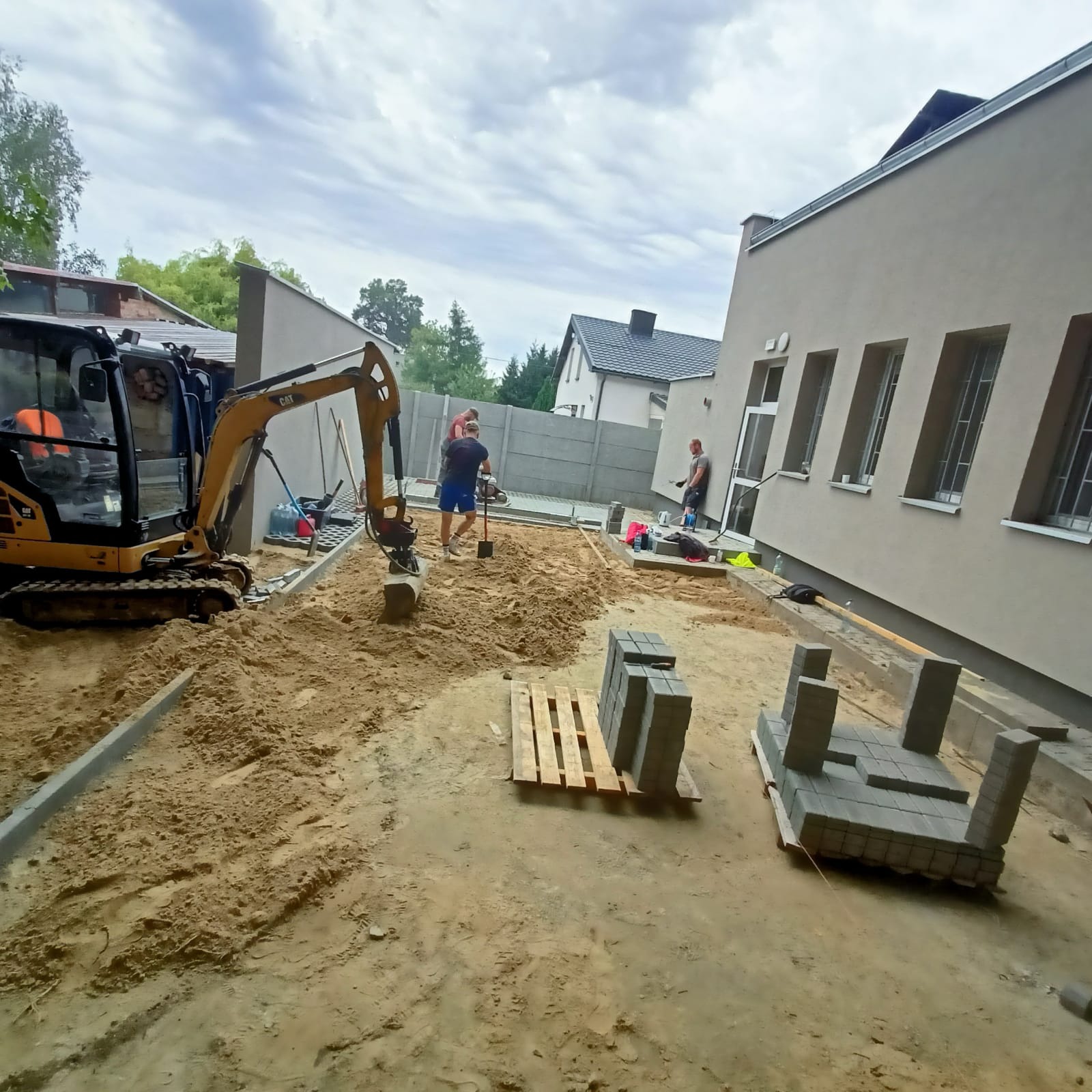 Żółta mini koparka CAT podczas prac ziemnych pod układanie kostki brukowej. Widoczni pracownicy i przygotowane kostki brukowe. Tło: budynek i ogrodzenie.