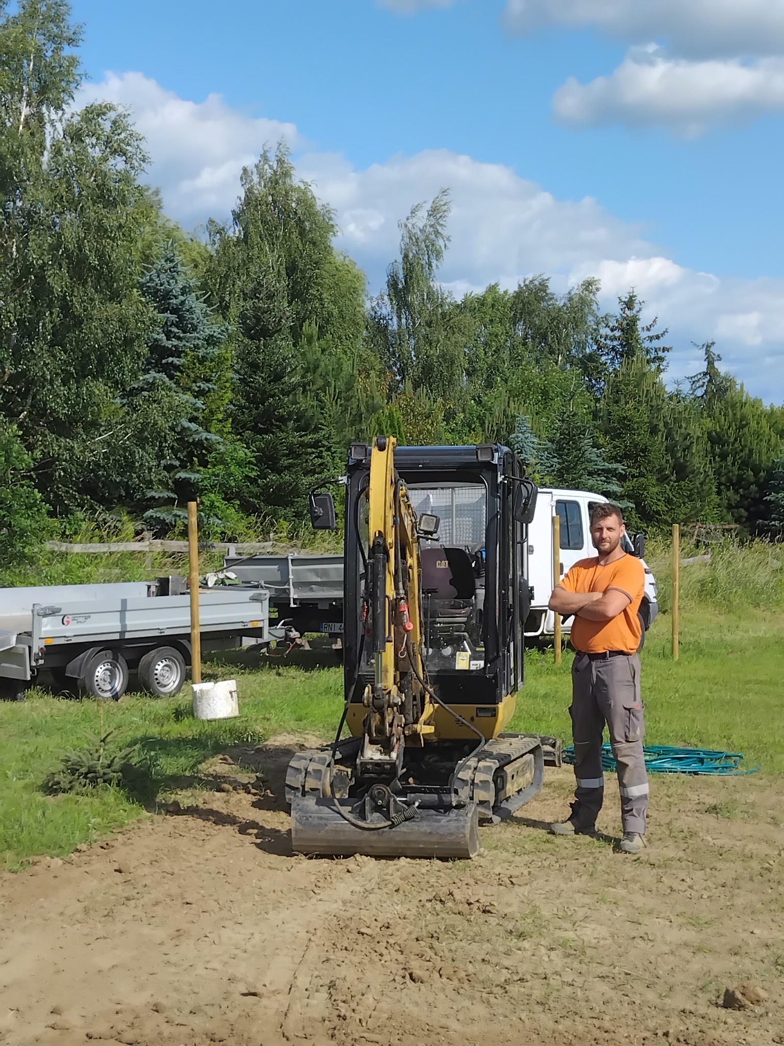 Żółta mini koparka CAT na placu budowy, obok stoi mężczyzna w pomarańczowej koszulce i szarych spodniach roboczych. W tle zieleń, przyczepa i biały bus.