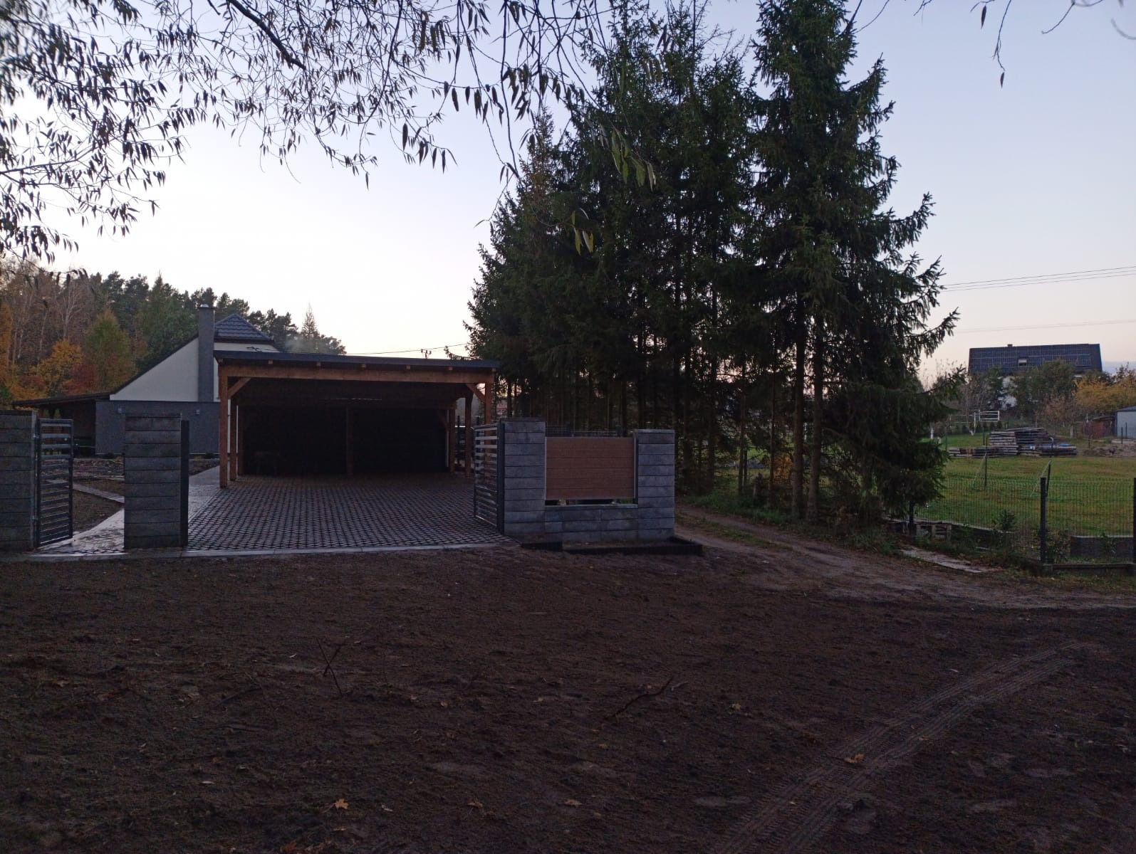 Ułożona kostka brukowa na podjeździe z wiatą, ogrodzenie z szarej kostki, przygotowany teren pod trawnik. Widok na dom i drzewa w tle, pochmurne niebo.