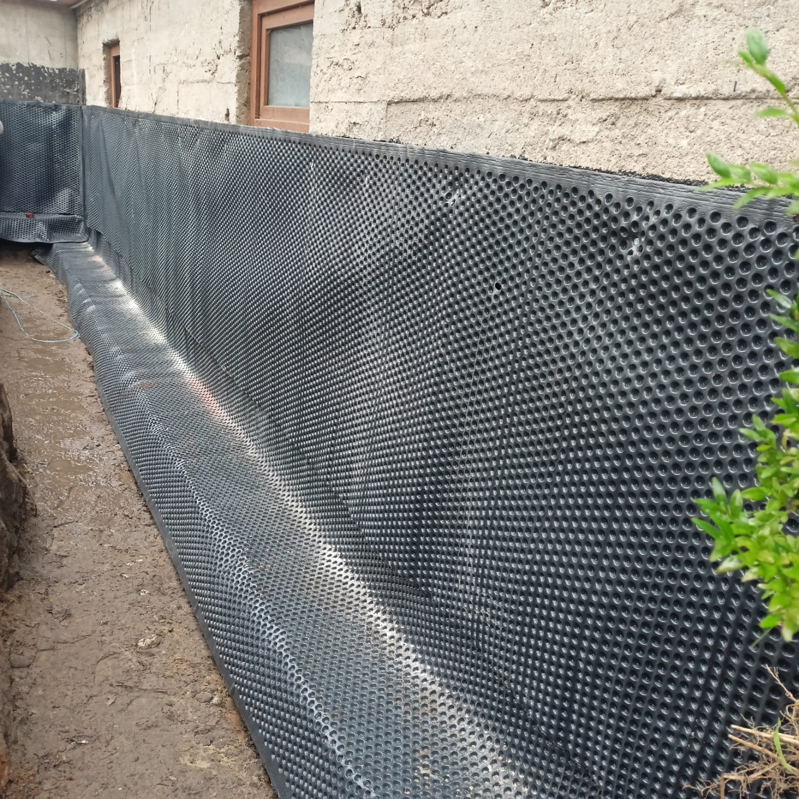 Czarna folia kubełkowa, ułożona w wykopie przy fundamencie budynku, zabezpieczająca przed wilgocią. Widoczny fragment wykopu i ściana budynku z oknem.