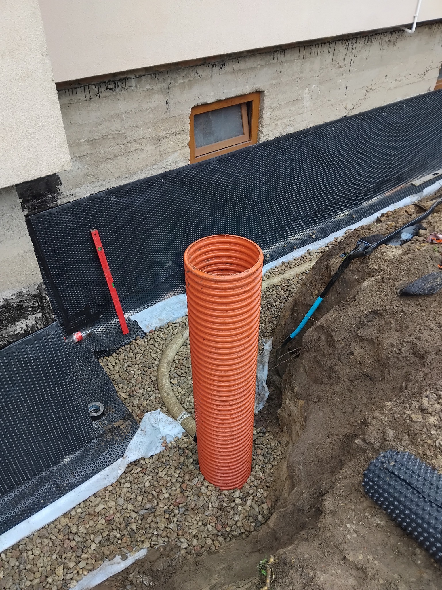 Fundament zabezpieczony folią kubełkową, obok rura drenarska w wykopie. Widoczne narzędzia i poziomica. Ochrona przed wilgocią w trakcie prac ziemnych.