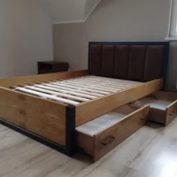 Drewniane łóżko z tapicerowanym zagłówkiem i szufladami, widoczne z boku, na podłodze. W tle stolik nocny i grzejnik.