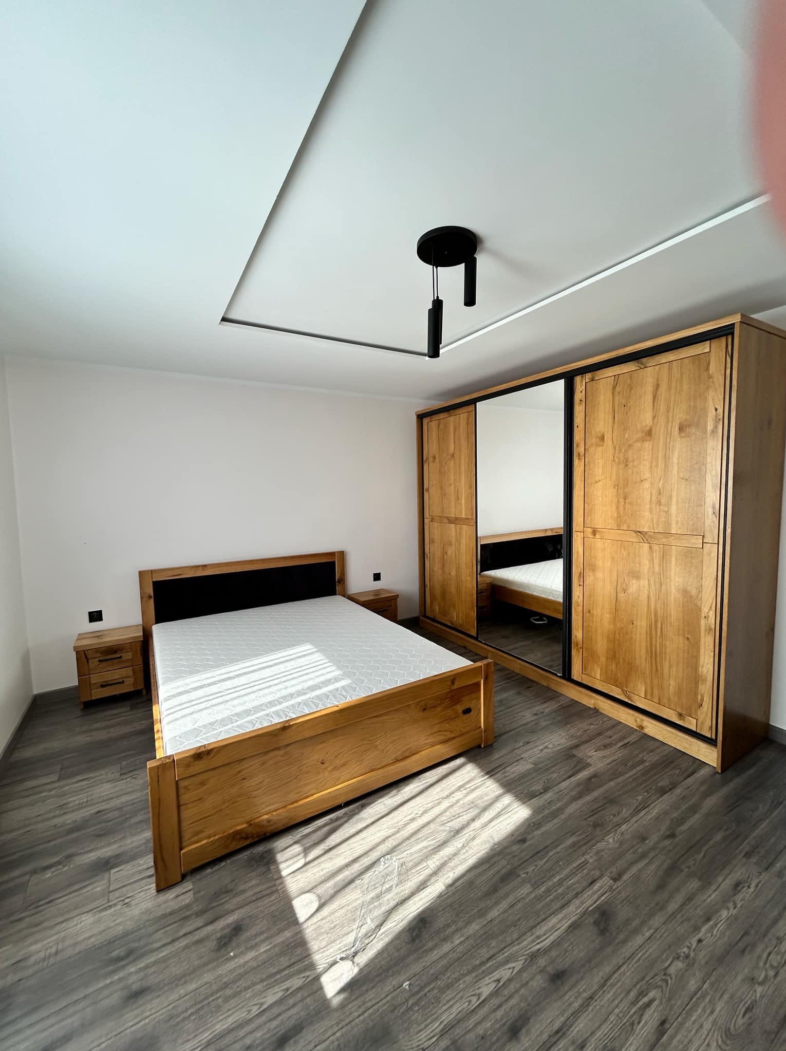 Drewniana szafa z lustrem przesuwnym, łóżko i szafki nocne w sypialni. Całość w ciepłym odcieniu drewna, na podłodze panele imitujące drewno.