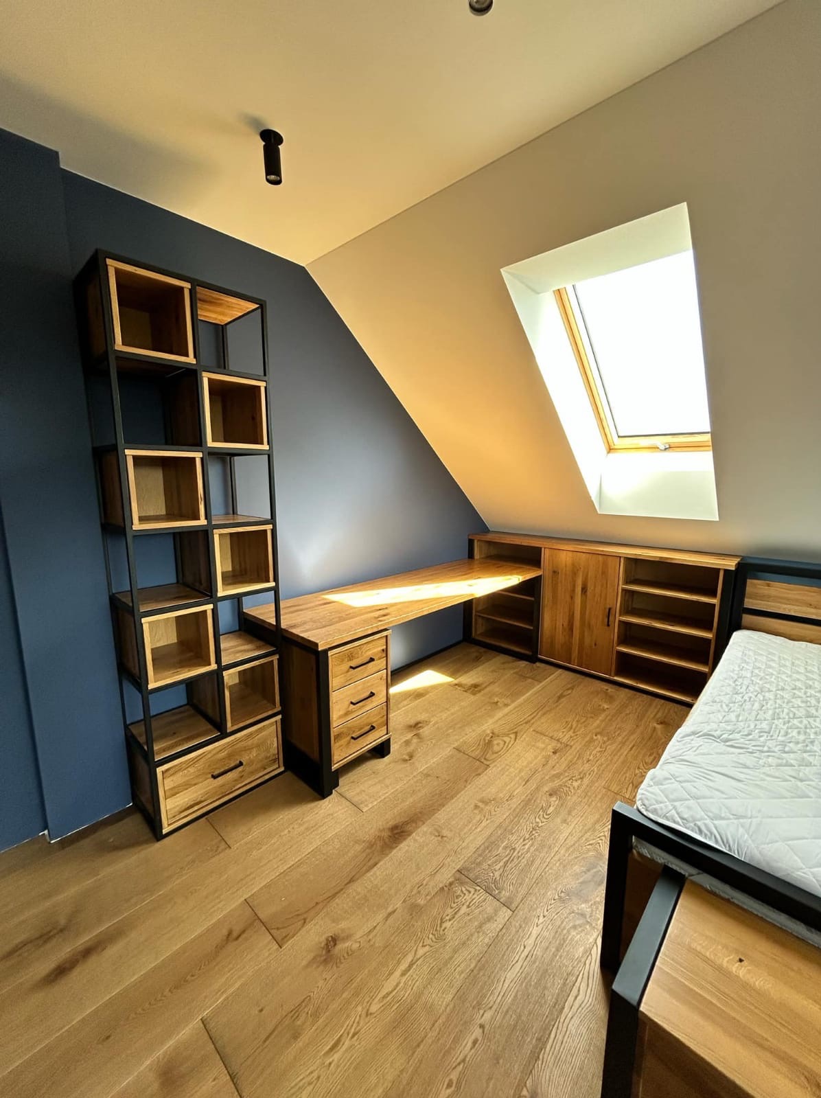 Pokój na poddaszu z drewnianym biurkiem, regałem i szafką w zabudowie. Czarna metalowa konstrukcja mebli kontrastuje z ciepłym odcieniem drewna i niebieską ścianą.