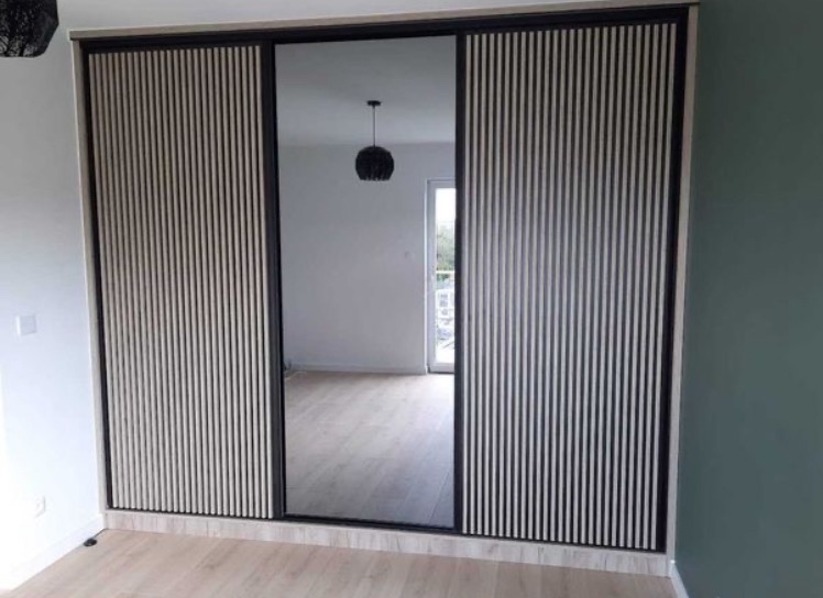 Nowoczesna szafa wnękowa z lustrem i lamelami na drzwiach przesuwnych. Minimalistyczny design, jasne drewno i czarne ramy. Widok na wnętrze pokoju z lampą.