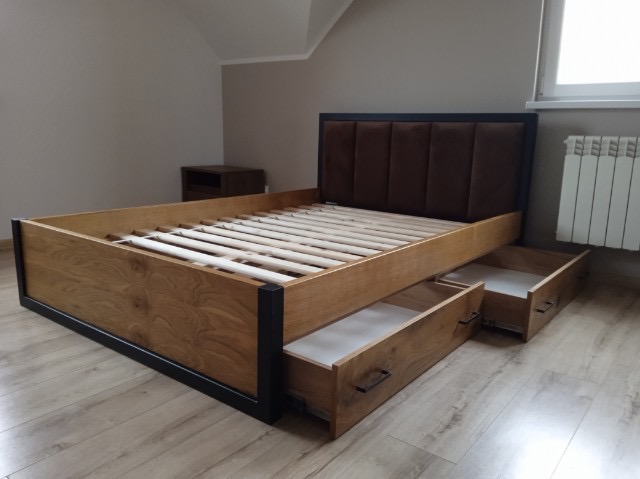 Drewniane łóżko z tapicerowanym zagłówkiem i szufladami, widoczne z boku, na podłodze. W tle stolik nocny i grzejnik.