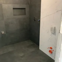 Wnętrze łazienki w trakcie wykańczania: szare, matowe płytki na ścianach i podłodze, wnęka w ścianie, biała ściana z marmurowym wzorem, widoczne instalacje hydrauliczne.