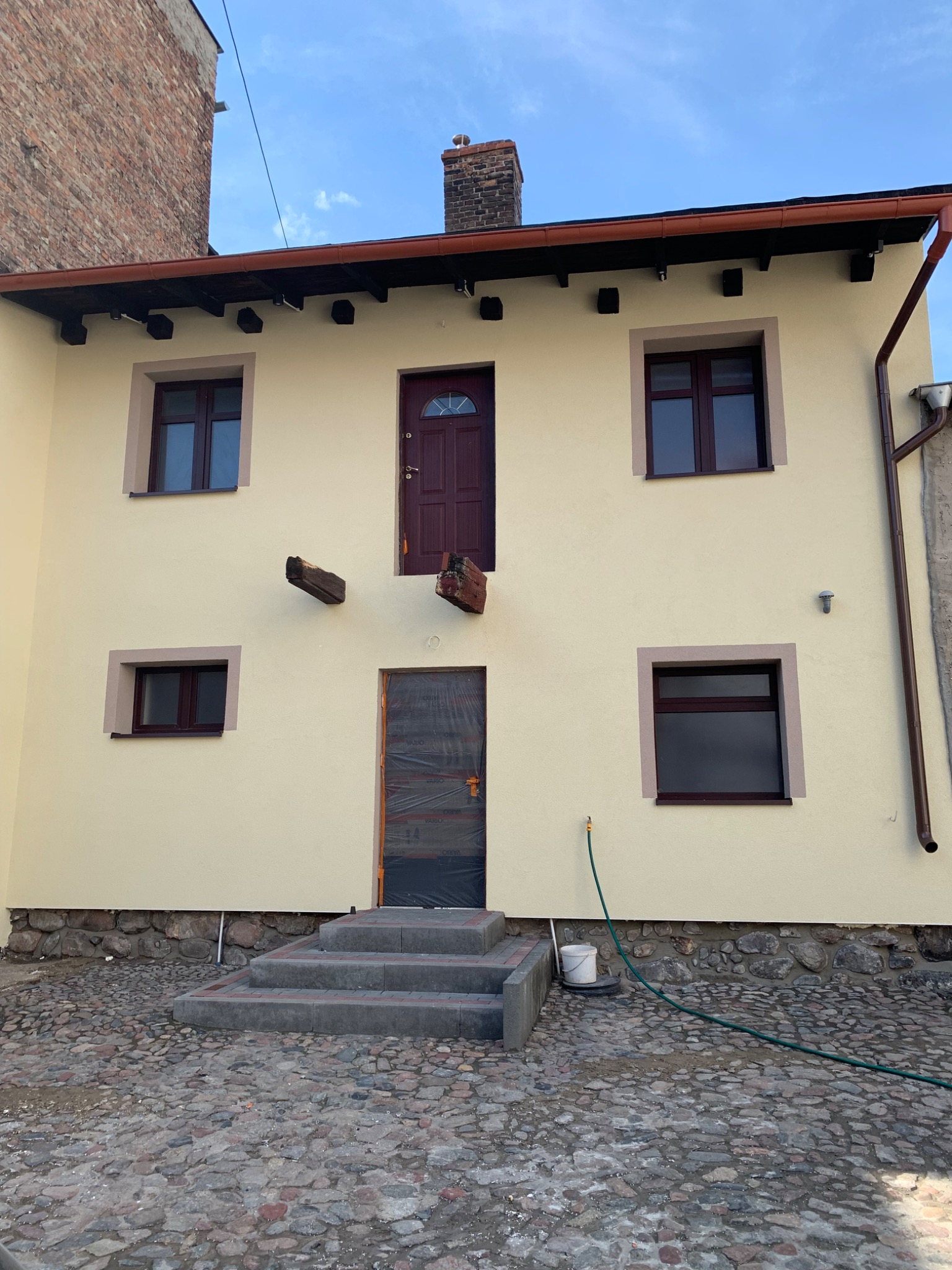 Odnowiona elewacja budynku z kremową farbą, nowymi oknami z brązowymi ramami i drewnianymi belkami dekoracyjnymi, schody wejściowe z kostki brukowej, widoczne zabezpieczenie drzwi folią malarską.