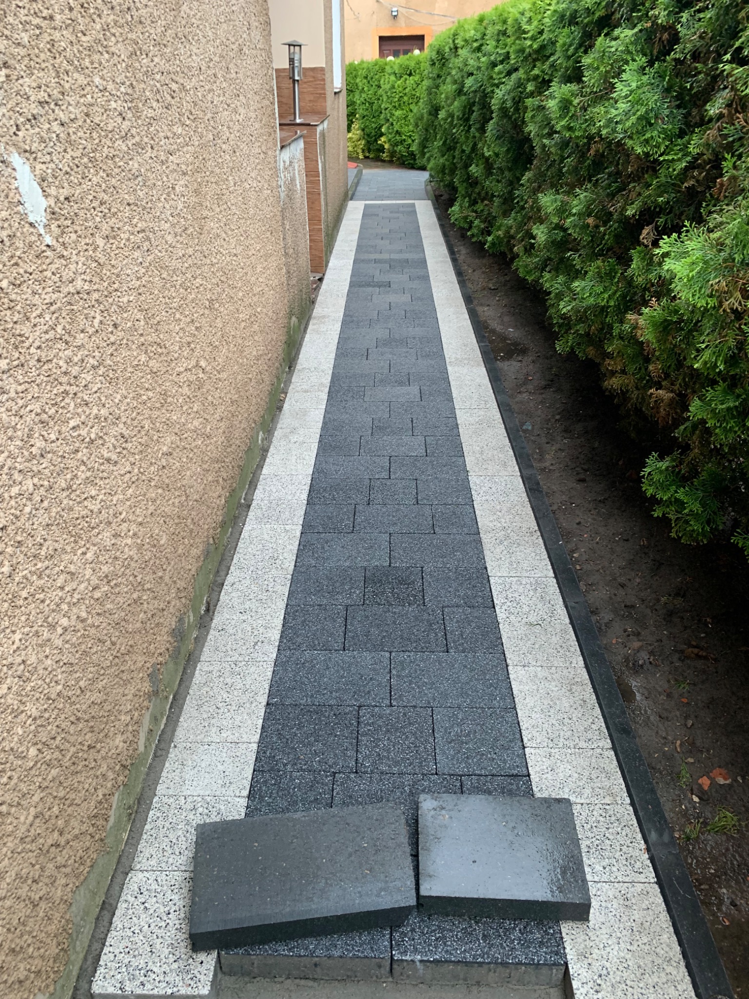 Nowo ułożony chodnik z kostki brukowej w odcieniach szarości i bieli, z dwoma luźnymi kostkami na wierzchu, biegnący wzdłuż ściany budynku i żywopłotu.
