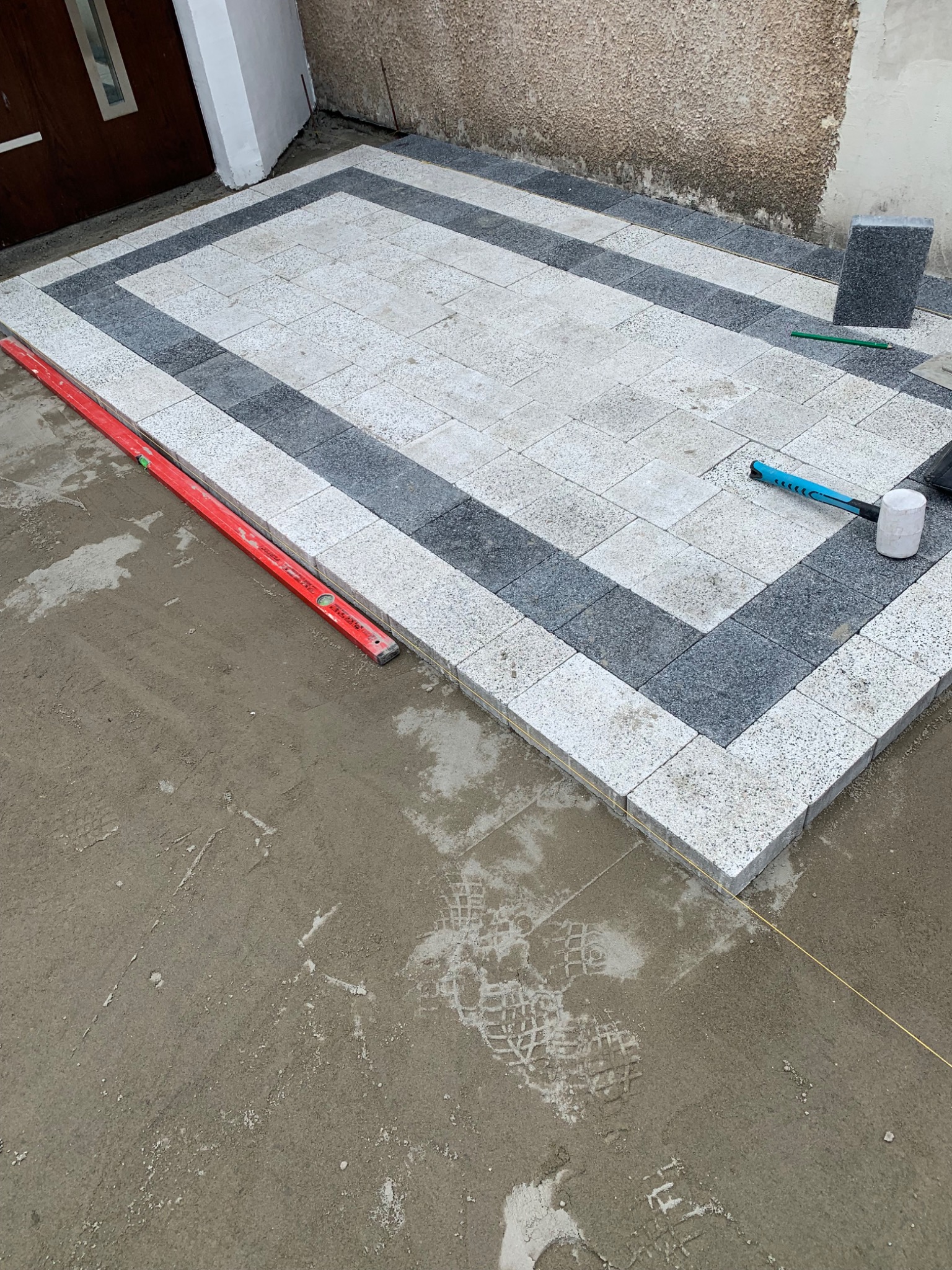 Układanie kostki brukowej w dwóch odcieniach szarości przed wejściem do domu, widoczna poziomica, gumowy młotek i zaprawa