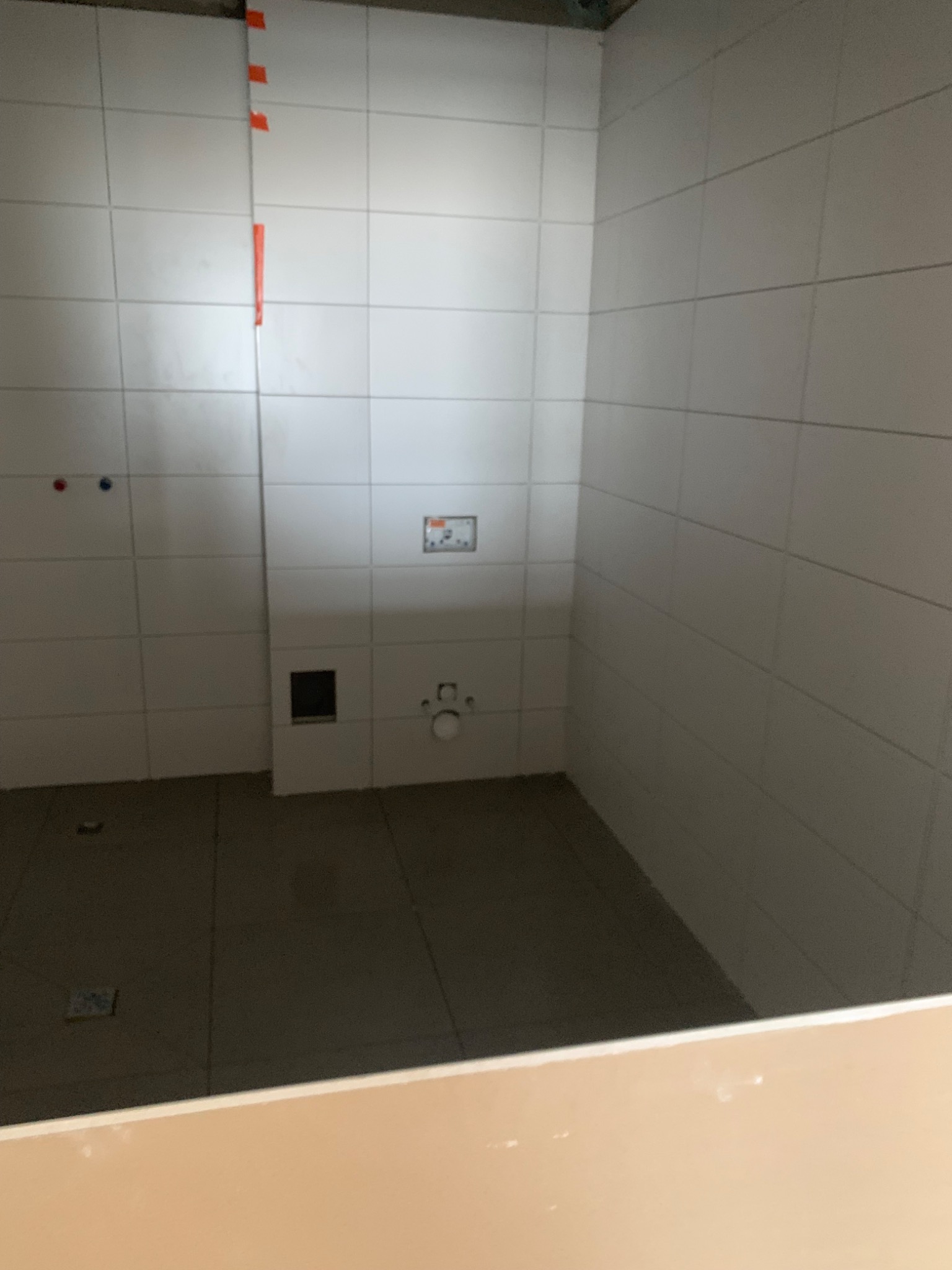 Wnętrze łazienki w trakcie układania białych, kwadratowych płytek, widoczne instalacje wodne i kanalizacyjne, zabezpieczone taśmą krawędzie płytek.