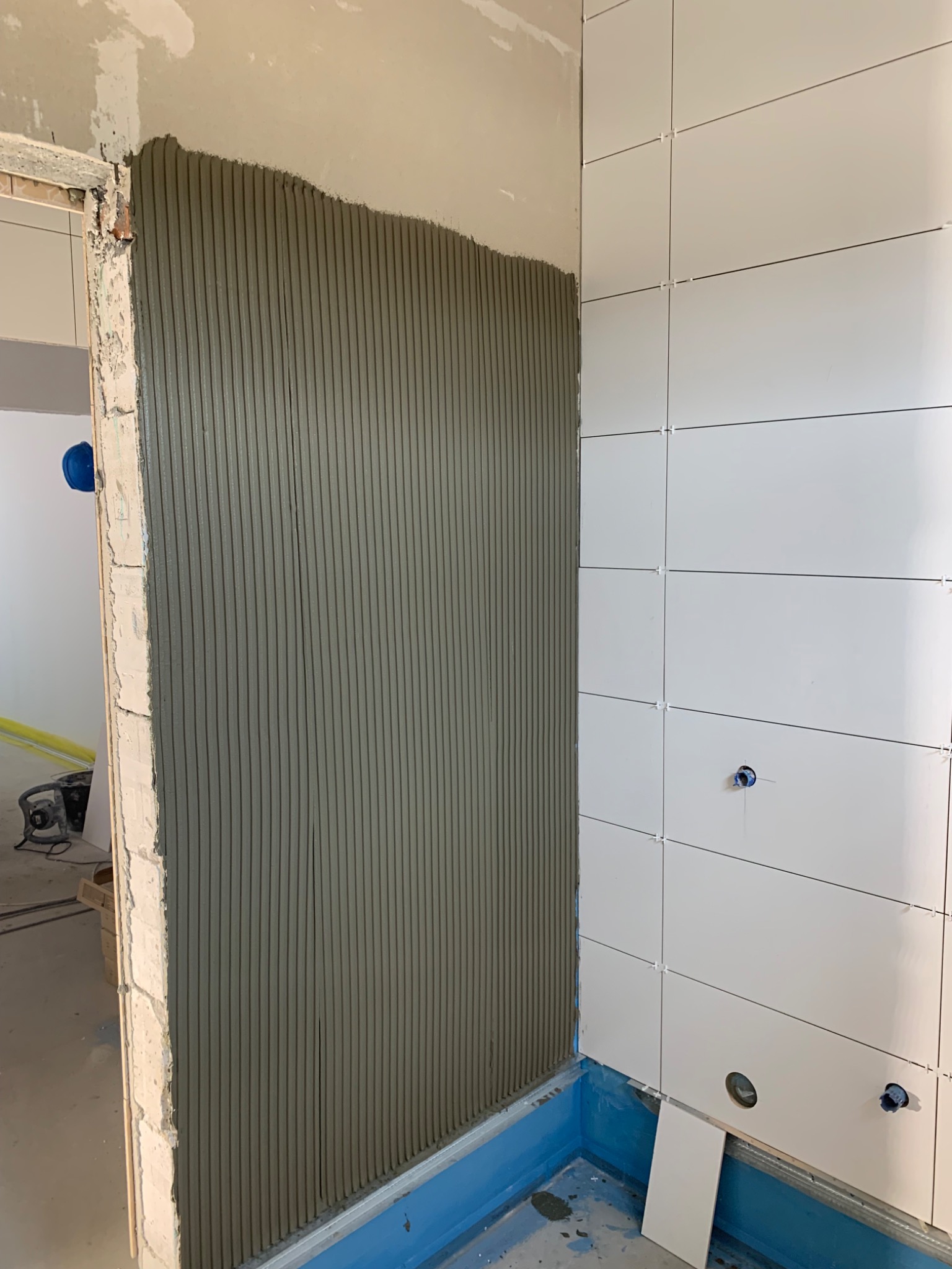 Ściana w trakcie kładzenia płytek, widoczna nałożona warstwa kleju w pionowe pasy oraz fragment ściany z ułożonymi białymi płytkami z krzyżykami dystansowymi w niebieskiej wannie.