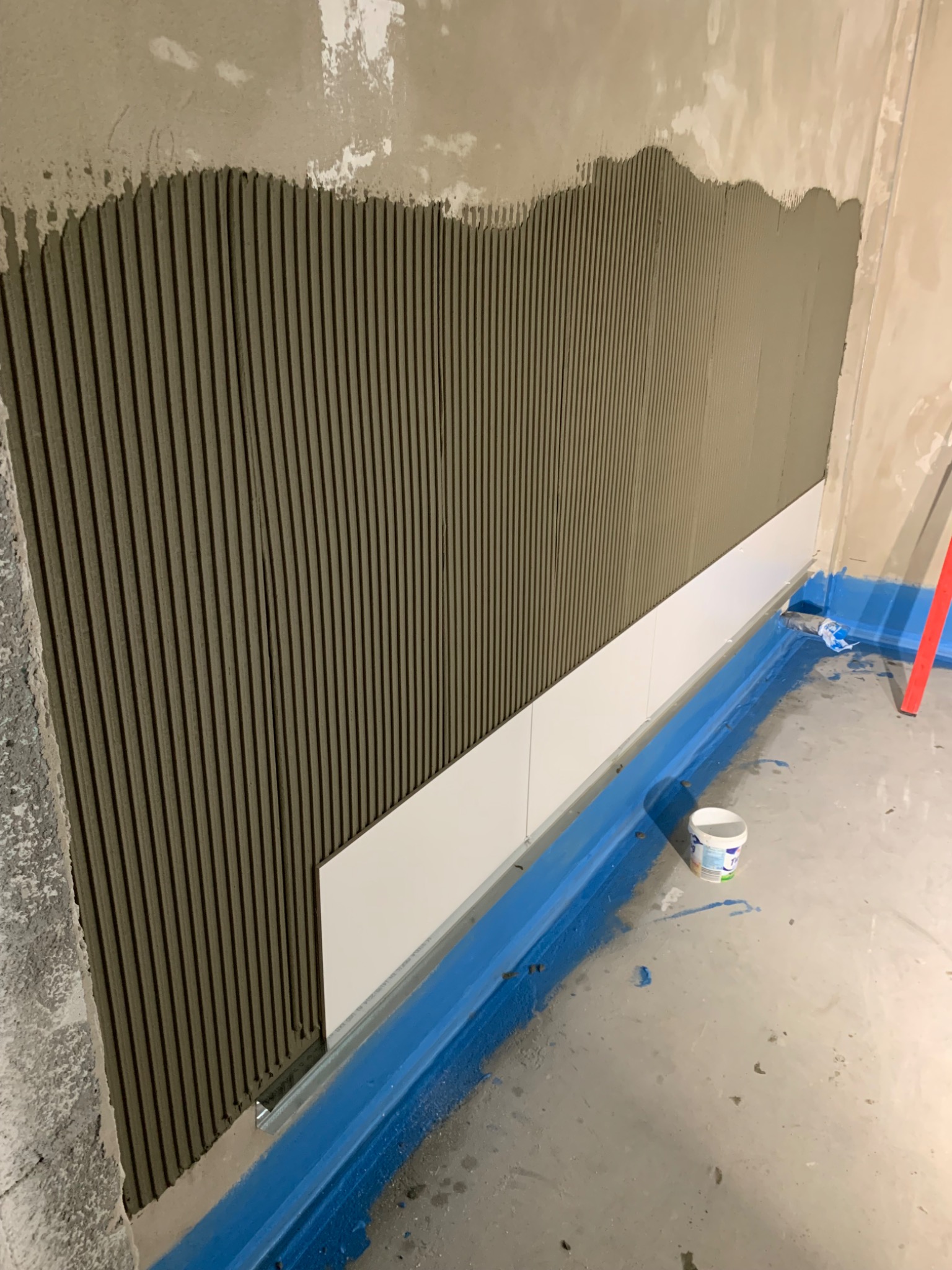 Ściana z nałożonym klejem w pionowe pasy, przygotowana do układania białych płytek, widoczna listwa poziomująca i zabezpieczenie podłogi folią w płynie.