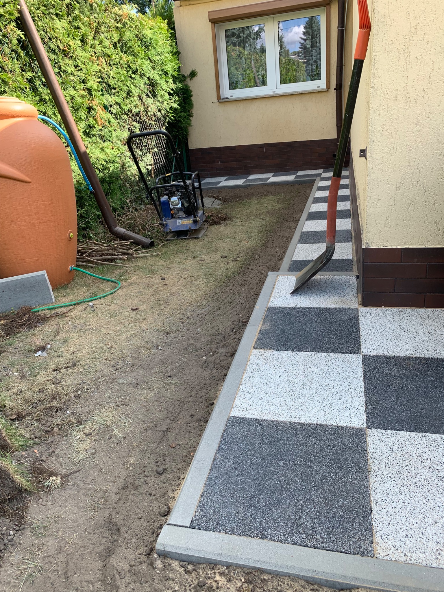 Ułożona szachownica z płyt chodnikowych w odcieniach szarości i bieli, obok ziemia przygotowana pod dalsze prace, widoczna ubijarka i zbiornik na deszczówkę.