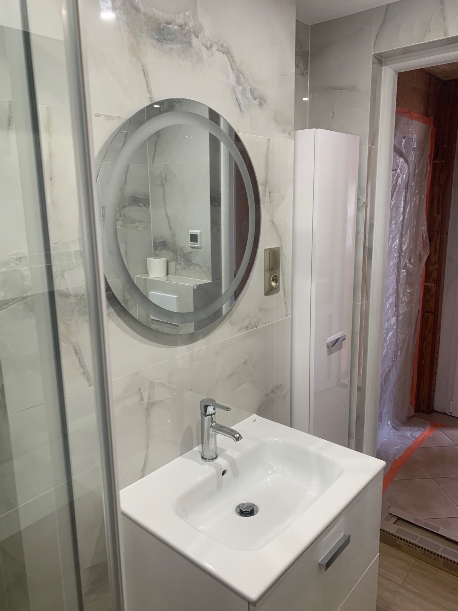 Biała umywalka z szafką i okrągłe lustro z podświetleniem w nowoczesnej łazience wykończonej marmurowymi płytkami. Widoczny fragment kabiny prysznicowej i zabezpieczone folią drzwi w trakcie remontu.