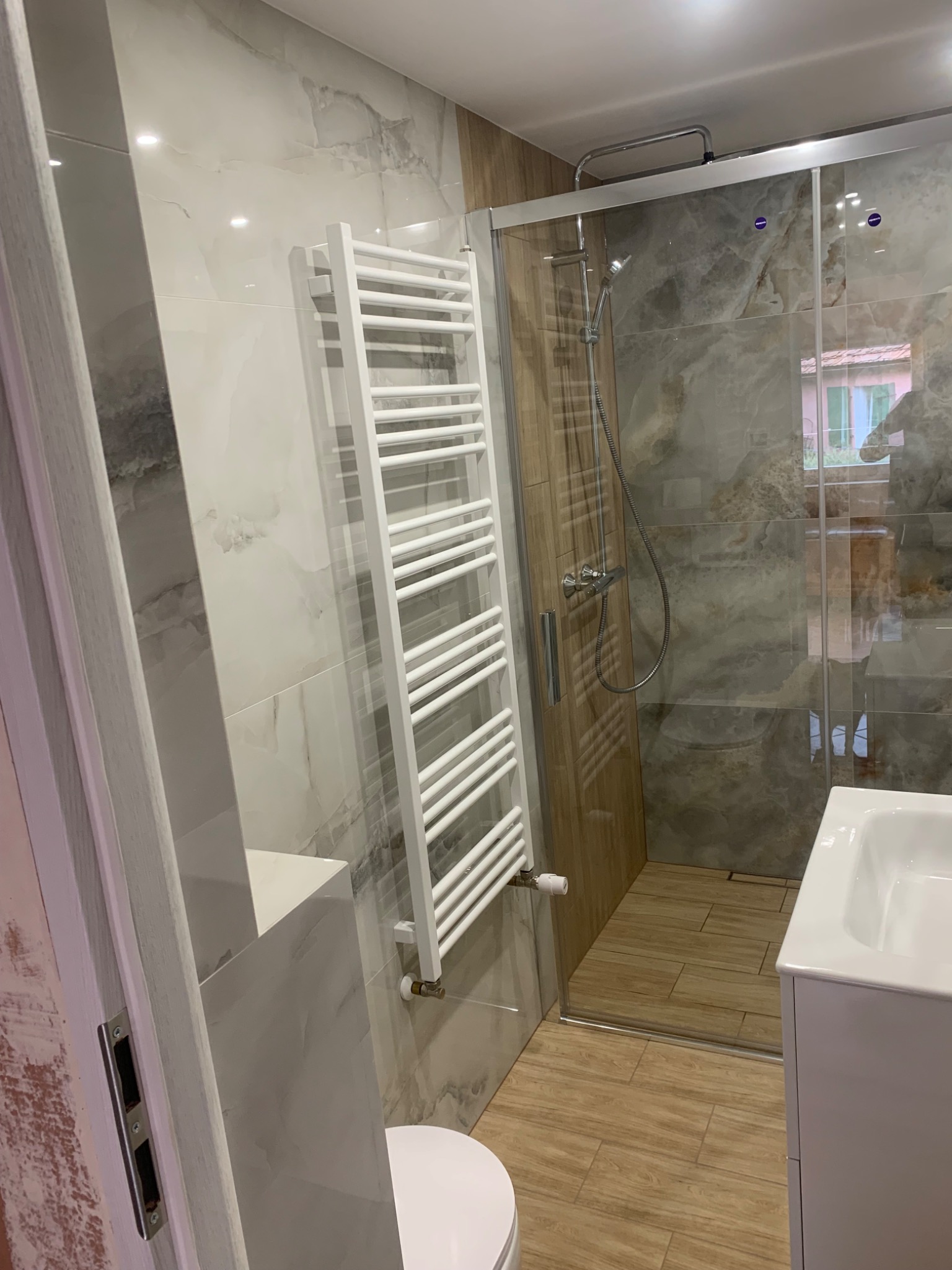 Wykończona łazienka z kabiną prysznicową, grzejnikiem drabinkowym i umywalką, wyłożona płytkami imitującymi marmur i drewno.