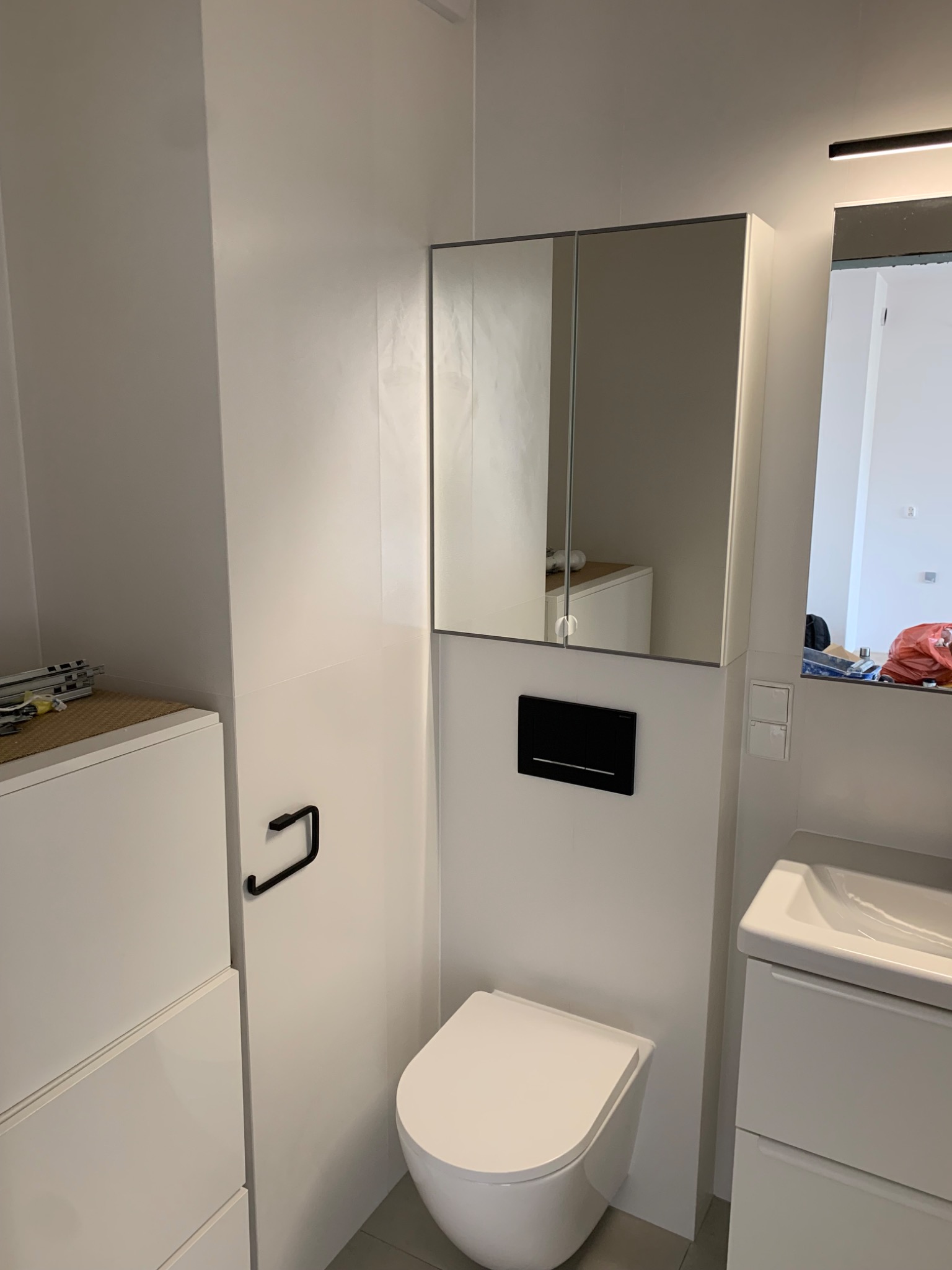 Nowoczesna, minimalistyczna łazienka z białą armaturą, podwieszaną toaletą z czarnym panelem sterującym i szafką z lustrem z oświetleniem LED, widoczne fragmenty umywalki i szafki pod umywalką.