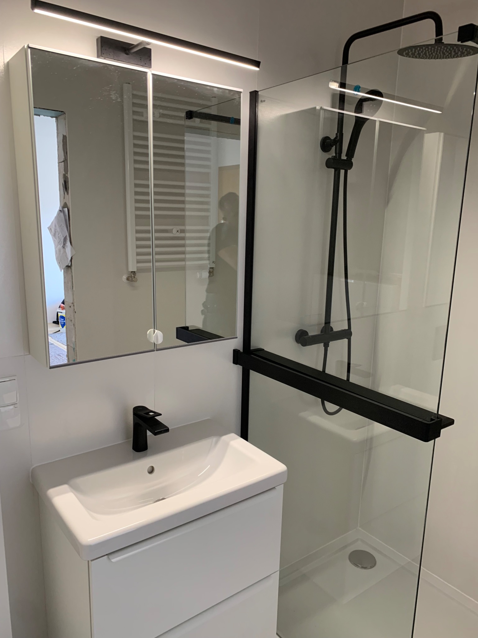 Nowoczesna łazienka z białą szafką pod umywalkę, czarną armaturą i szklaną kabiną prysznicową z czarnym wykończeniem, oświetlenie LED nad lustrem.