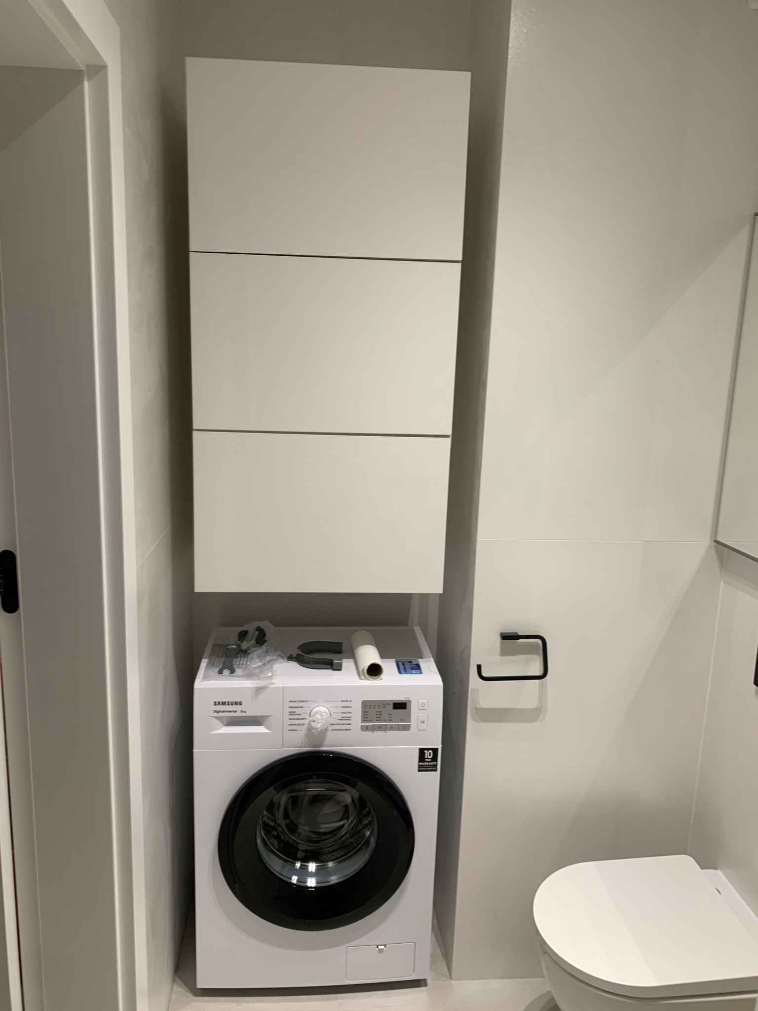 Nowoczesna, minimalistyczna łazienka z pralką Samsung, białą zabudową meblową i wiszącą toaletą. Na pralce widoczne akcesoria montażowe i instrukcja.