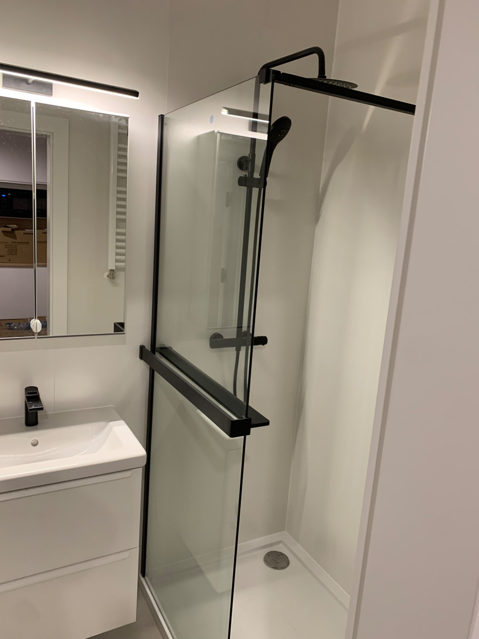 Nowoczesna kabina prysznicowa walk-in z czarnymi elementami, umywalka z szafką w minimalistycznej łazience, widoczne białe ściany i oświetlenie LED nad lustrem.