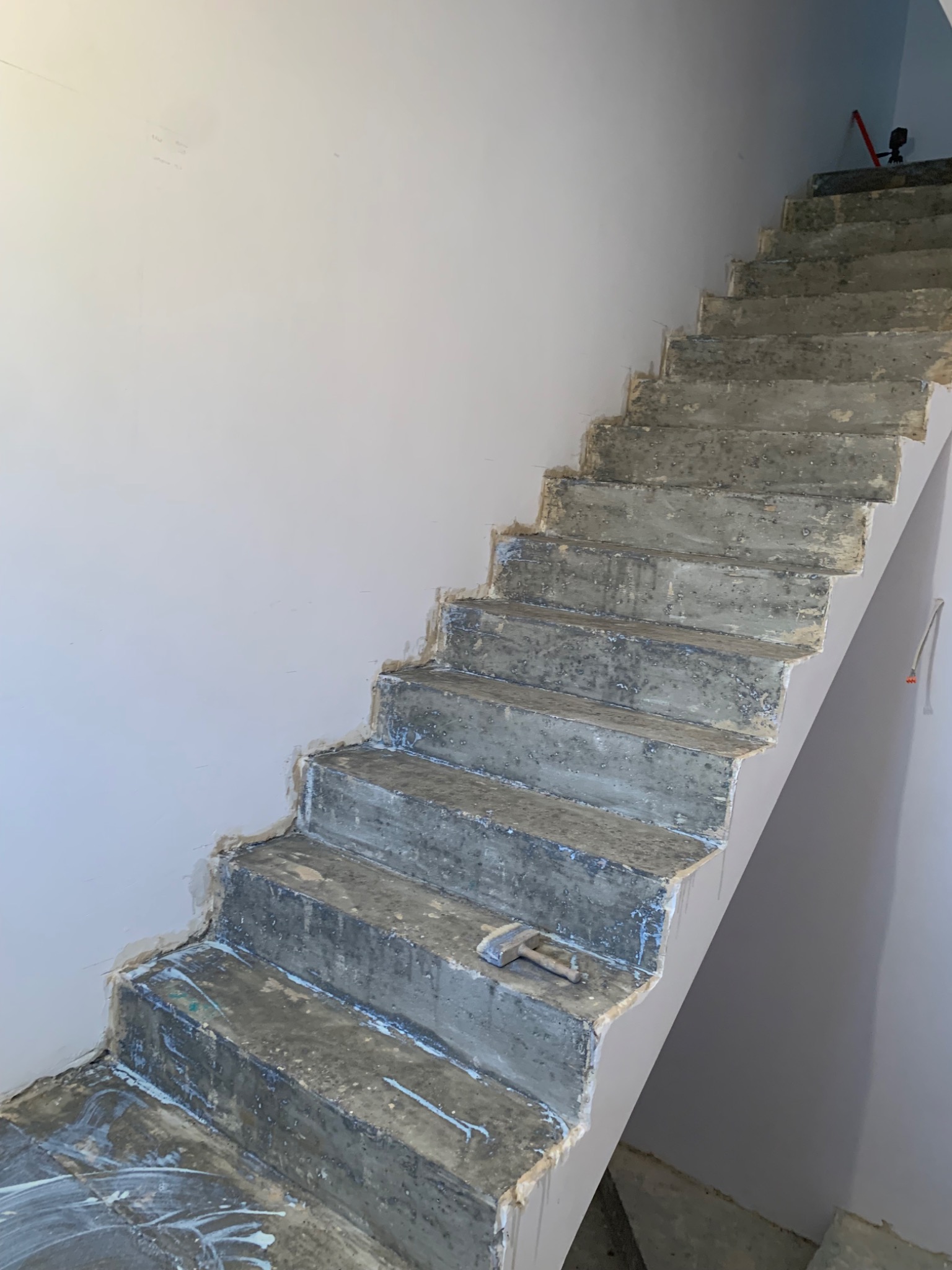 Betonowe schody w trakcie prac remontowych, widoczne ślady zaprawy i młotek na jednym ze stopni, ściany przygotowane do malowania.