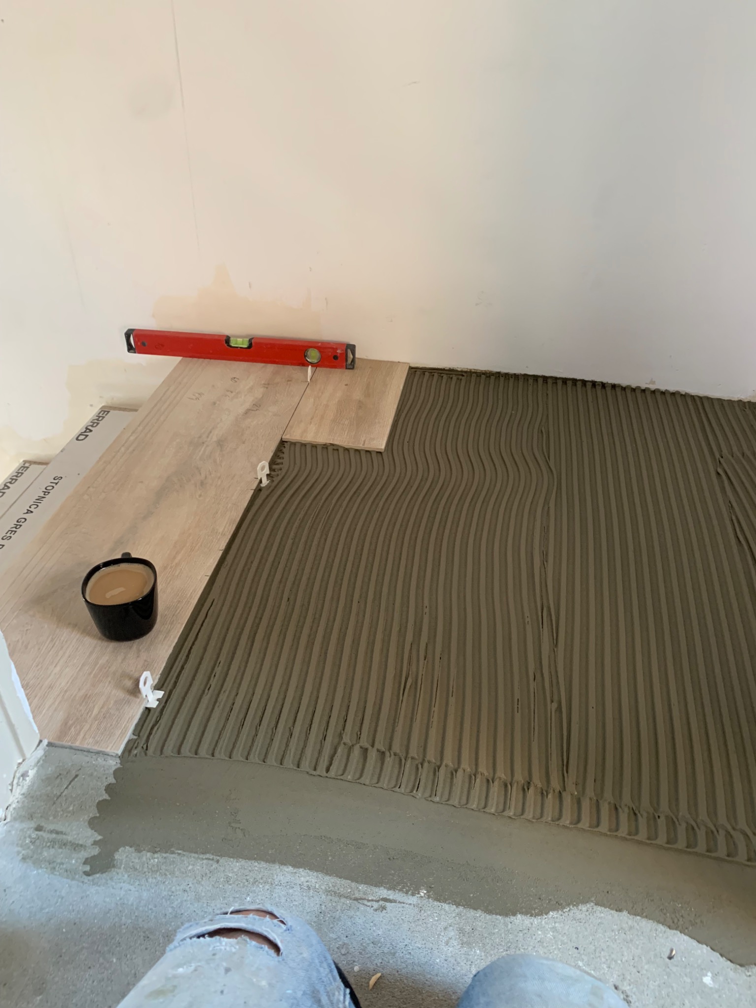 Układanie jasnych płytek podłogowych z użyciem poziomicy i krzyżyków dystansowych, widoczna filiżanka z kawą i spodnie robocze w dolnej części kadru.