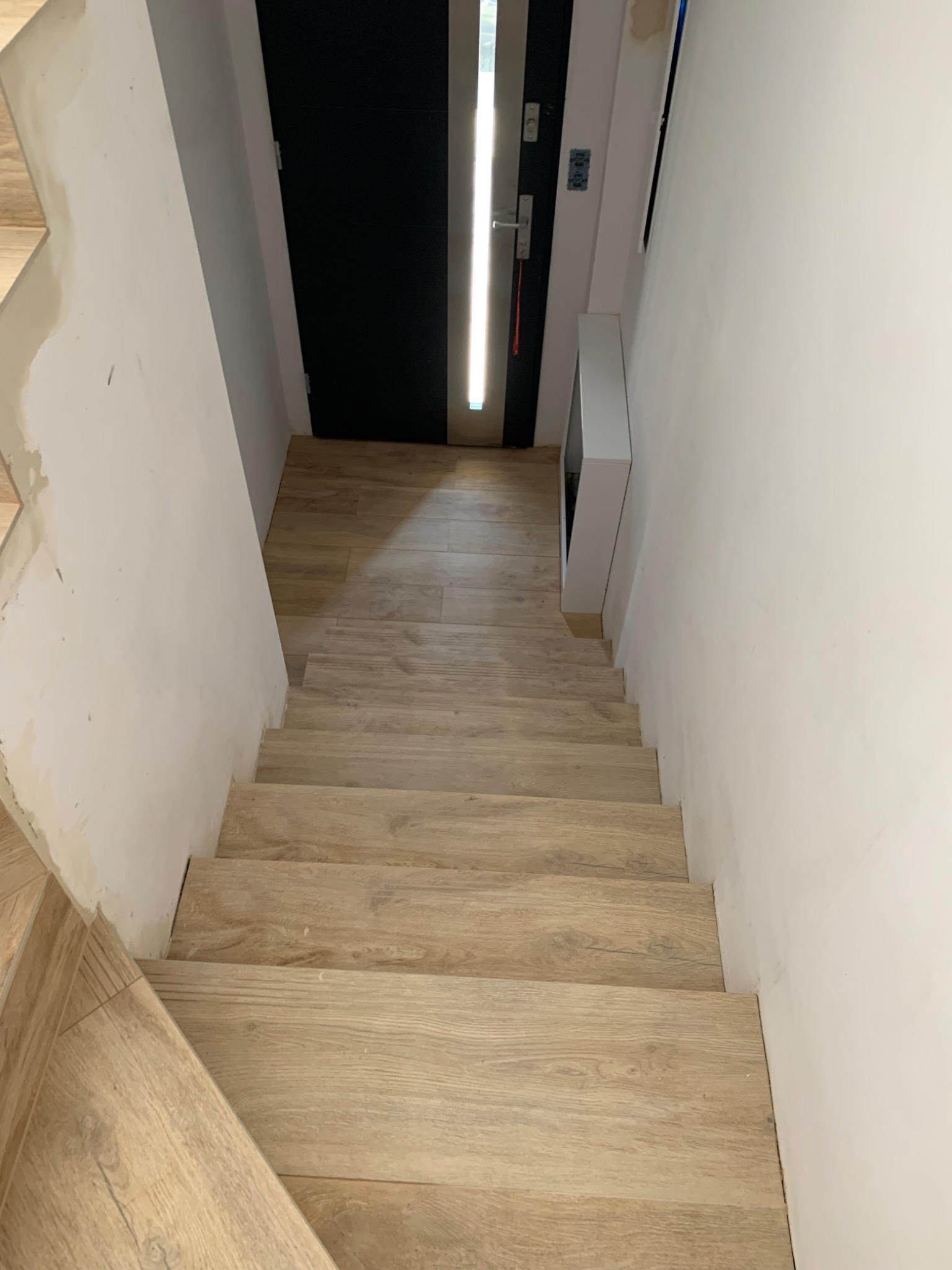 Wykończone schody z panelami imitującymi drewno prowadzące do czarnych drzwi, widok z góry na niepomalowaną ścianę z widocznymi śladami po szpachlowaniu.