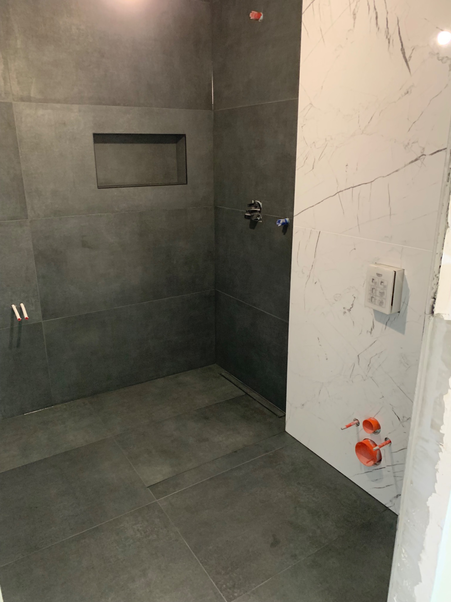 Wnętrze łazienki w trakcie wykańczania: szare, matowe płytki na ścianach i podłodze, wnęka w ścianie, biała ściana z marmurowym wzorem, widoczne instalacje hydrauliczne.