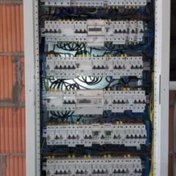 Rozdzielnia elektryczna z zabezpieczeniami, licznikami i przewodami, umieszczona w metalowej obudowie na tle ściany z cegły.