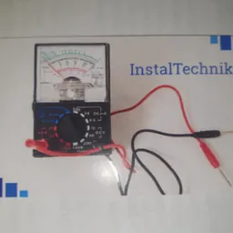 Analogowy multimetr z przewodami pomiarowymi na tle z logo InstalTechnik, widok z góry.