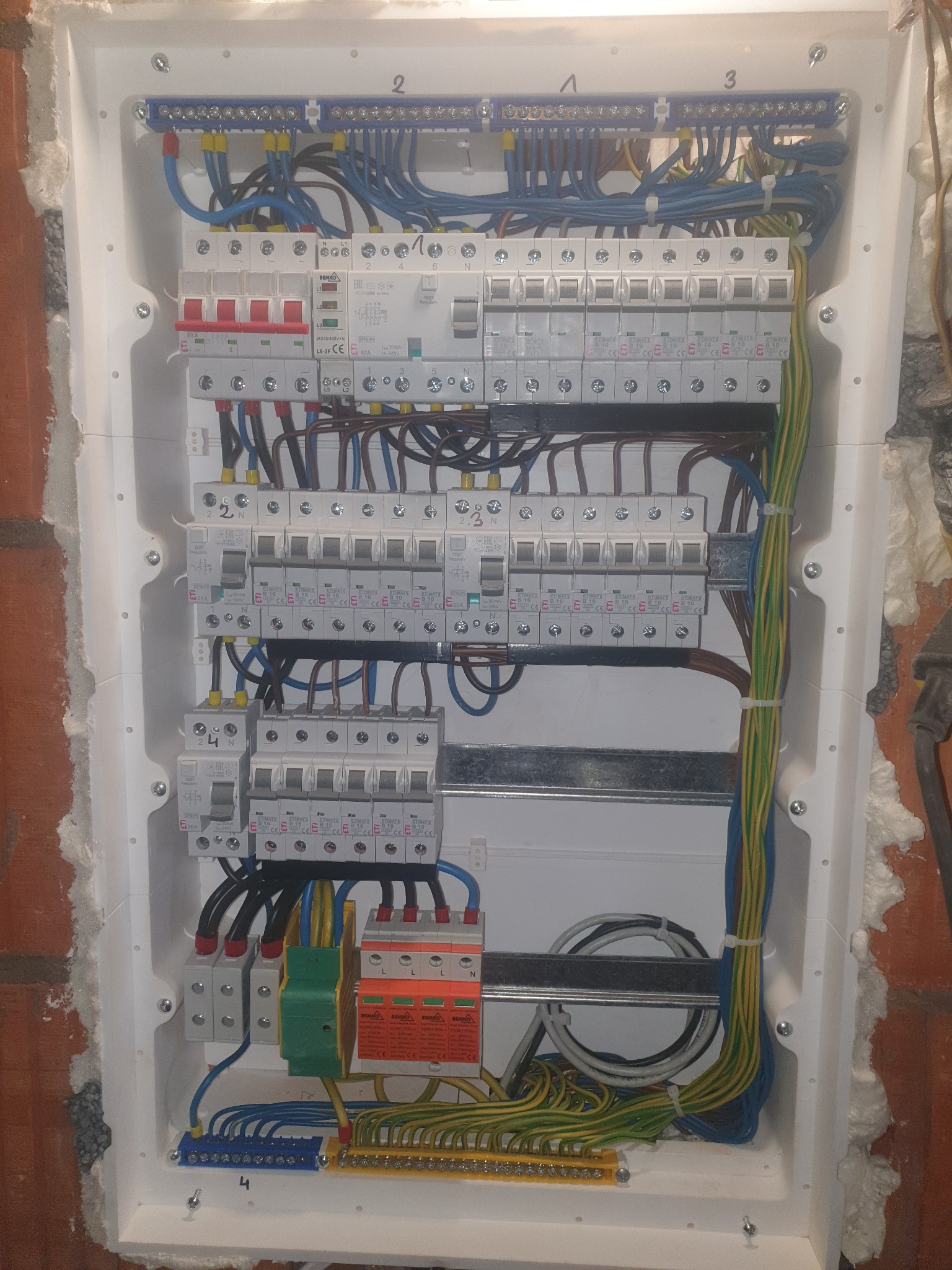 Rozdzielnia elektryczna w białej obudowie, widoczne zabezpieczenia różnicowoprądowe, wyłączniki nadprądowe, listwy zaciskowe i przewody w różnych kolorach, zamontowana w ścianie z czerwonej cegły...