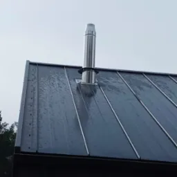 Błyszczący, stalowy komin wystający z mokrego, ciemnego dachu blaszanego w pochmurny dzień.