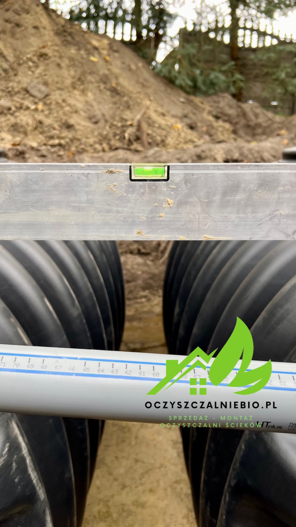 Montaż rur kanalizacyjnych z użyciem poziomicy, widoczny wykop ziemny w tle. Na pierwszym planie rura z logo firmy zajmującej się sprzedażą i montażem oczyszczalni ścieków.