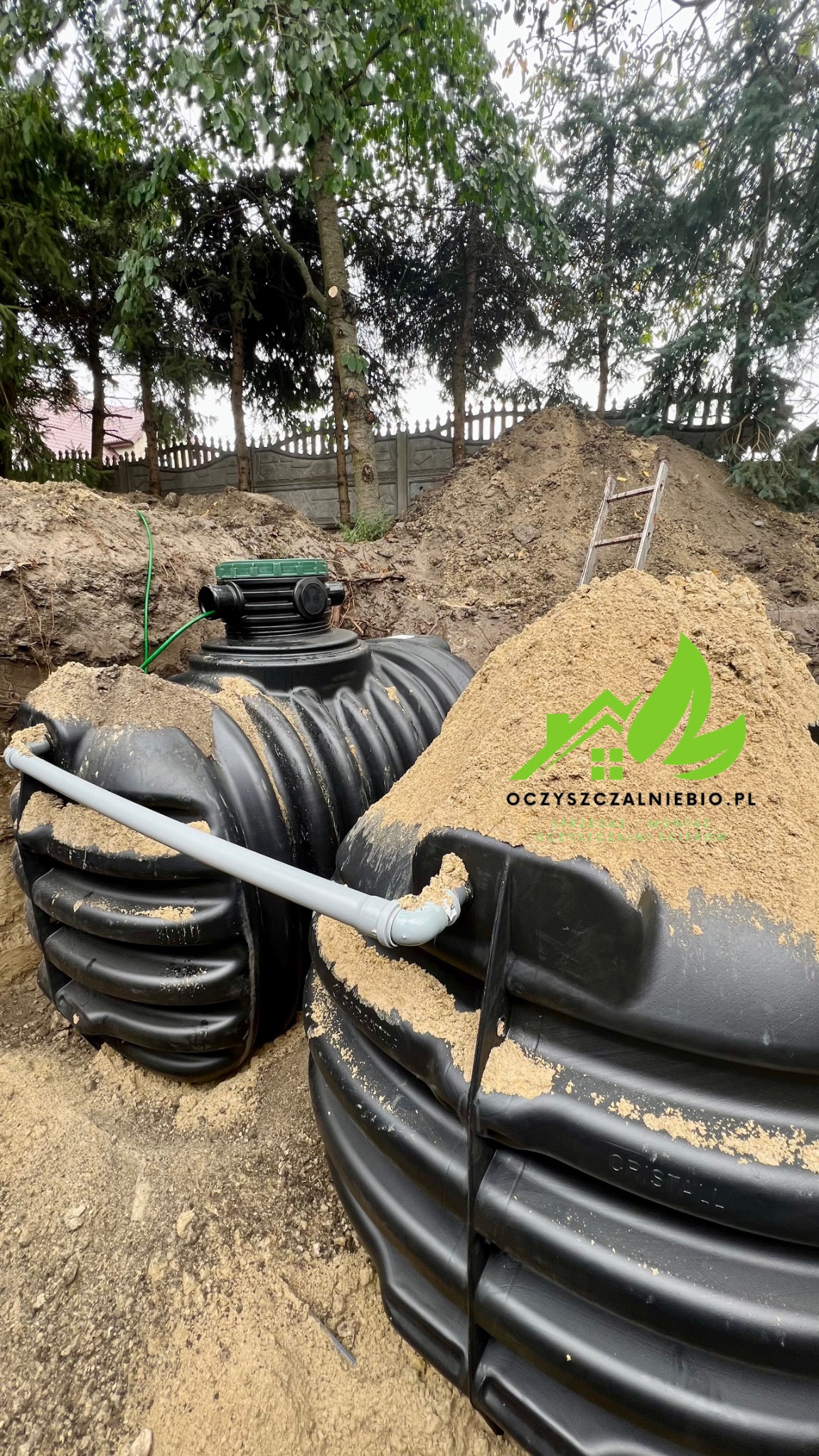 Dwa zbiorniki z tworzywa sztucznego, częściowo zasypane piaskiem, połączone rurą PCV na tle wykopu i drabiny opierającej się o pryzmę ziemi.