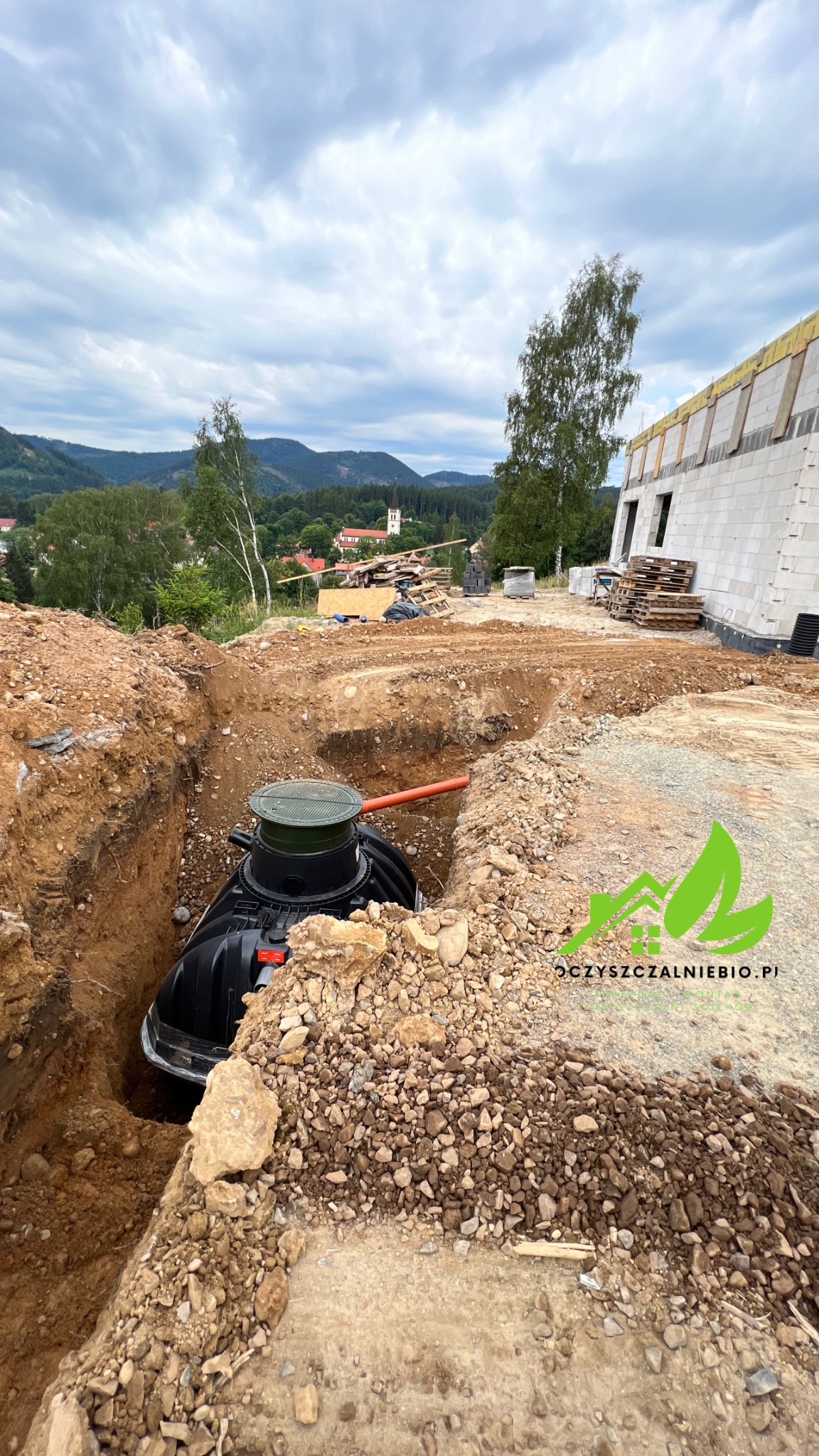 Czarna, plastikowa oczyszczalnia w wykopie ziemnym, z widocznym pomarańczowym rurociągiem, w tle budowa domu jednorodzinnego i krajobraz wiejski z kościołem.
