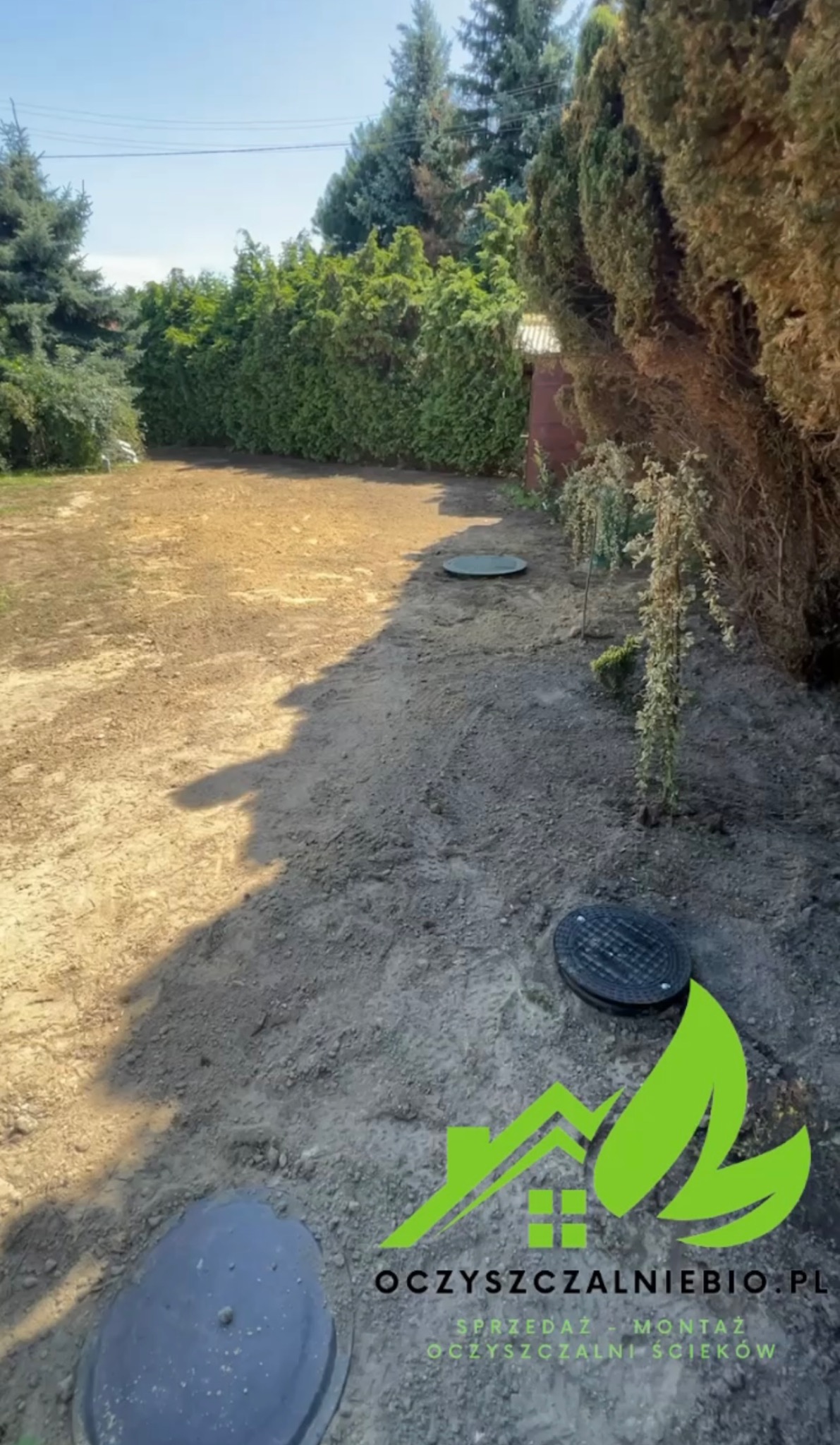 Teren po instalacji przydomowej oczyszczalni ścieków z widocznymi włazami inspekcyjnymi, świeżo zasypany ziemią, z logotypem firmy w prawym dolnym rogu.