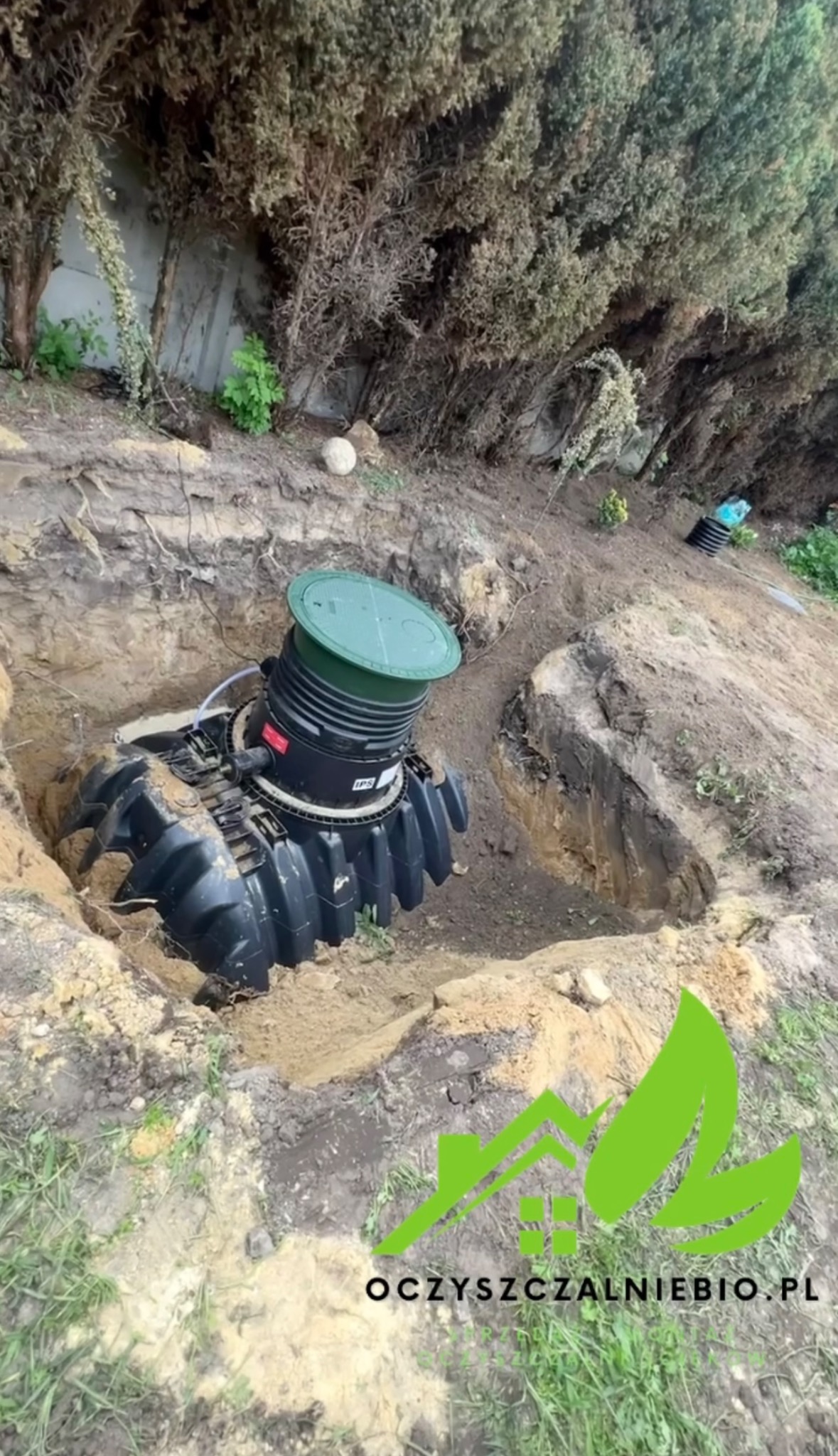 Montaż czarnej, segmentowej konstrukcji z zieloną pokrywą w wykopie ziemnym, widoczne elementy instalacji i ziemia wokół, w tle żywopłot.