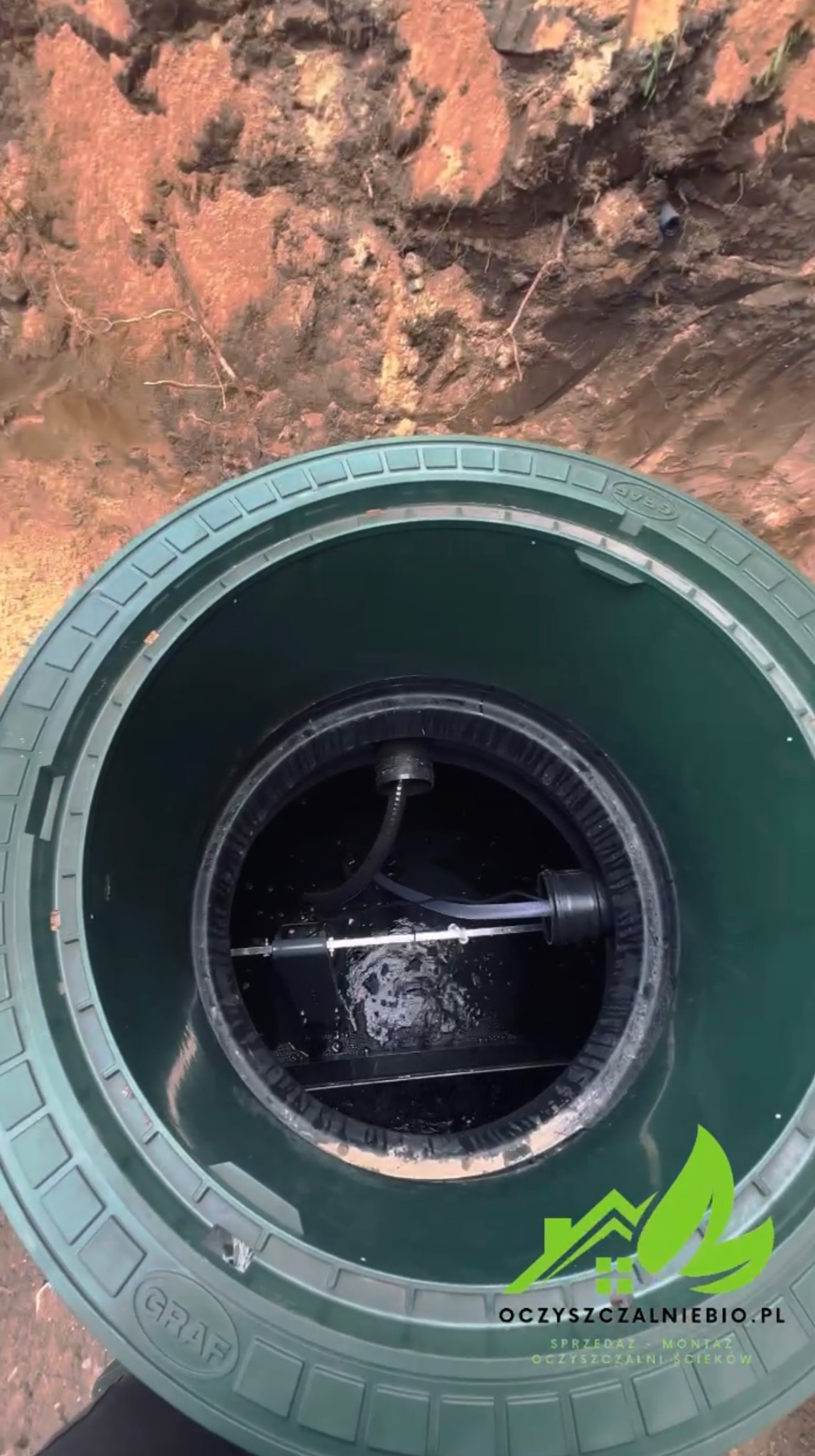 Otwarty właz przydomowej oczyszczalni ścieków Graf, widok z góry na wnętrze zbiornika z elementami instalacji, ziemia w tle.