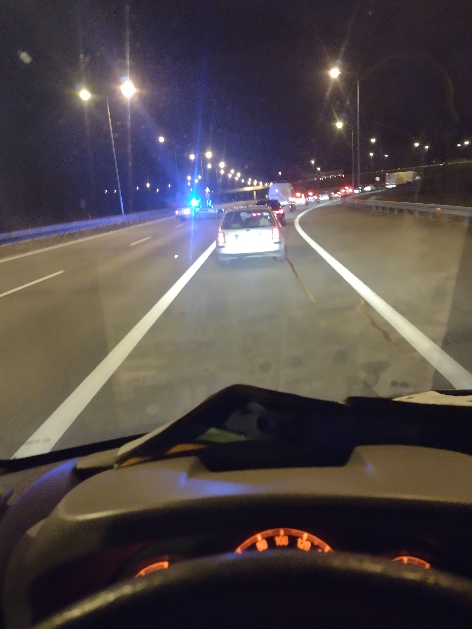 Widok z kabiny kierowcy na nocną autostradę z widocznymi światłami samochodów i latarni ulicznych; w oddali światła alarmowe.
