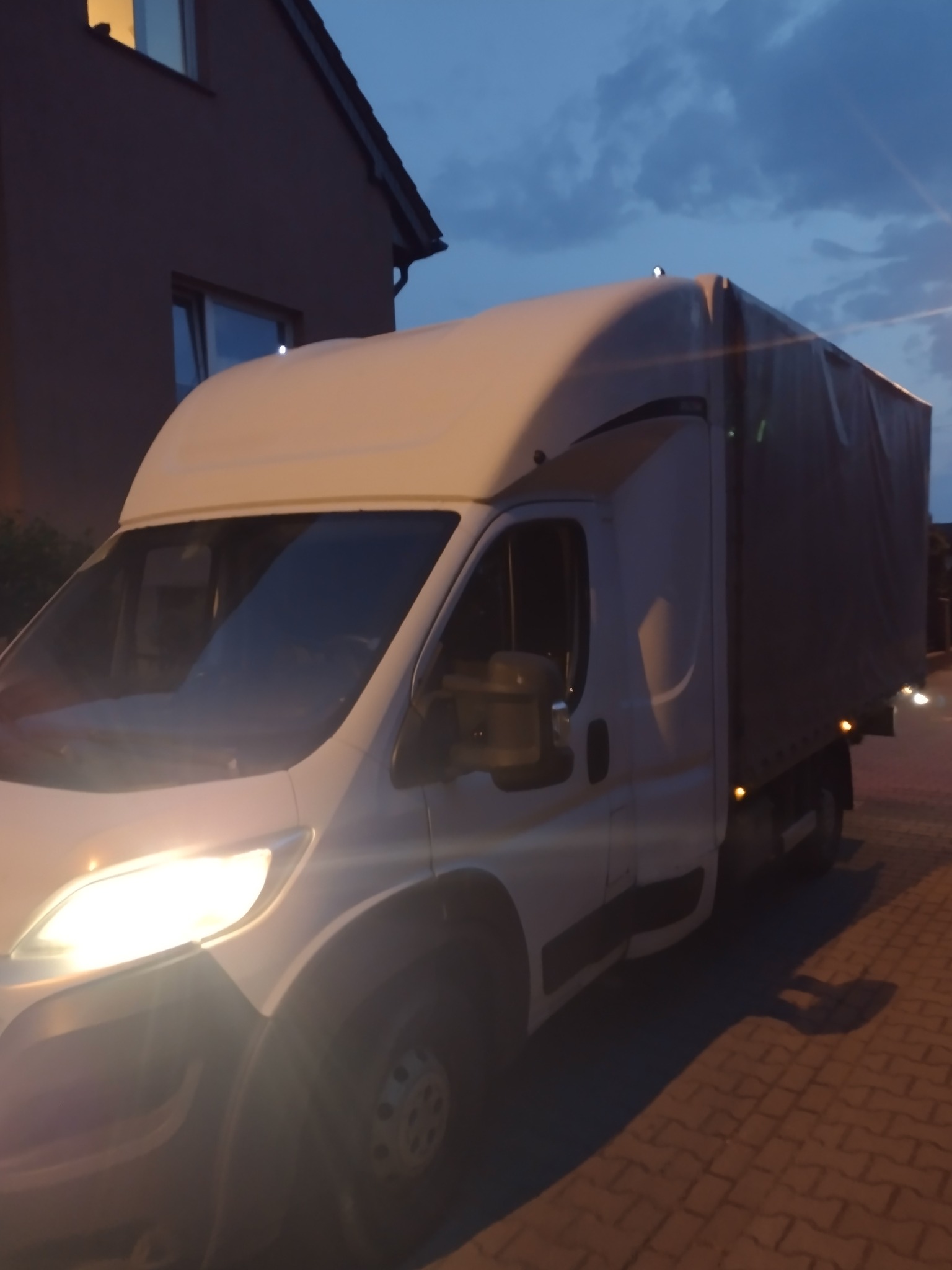 Biały samochód dostawczy z plandeką zaparkowany na brukowanej ulicy o zmierzchu, widoczne światła przednie i fragment budynku w tle.