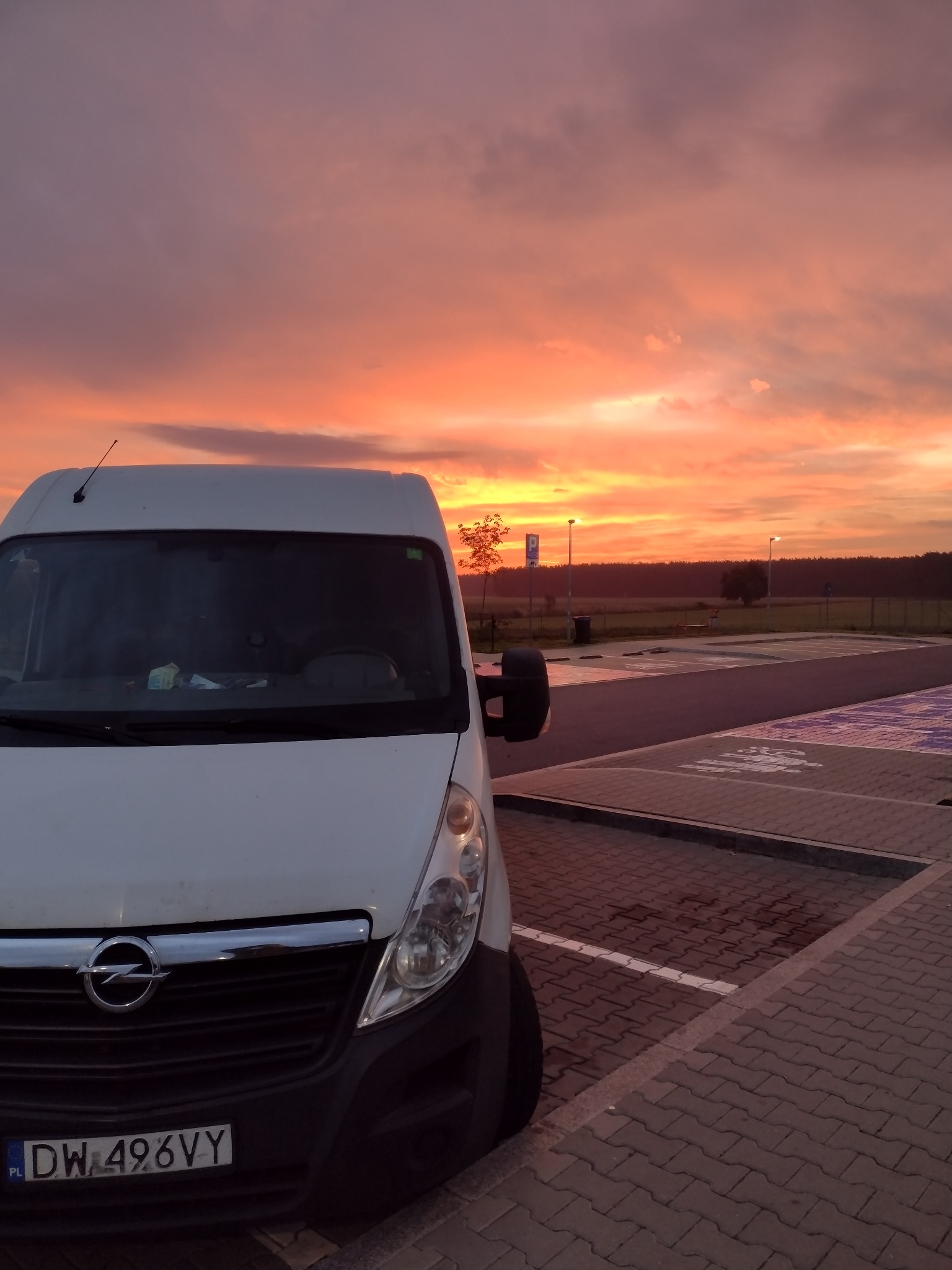 Biały bus marki Opel zaparkowany na parkingu z kostki brukowej o zachodzie słońca, widoczna tablica rejestracyjna DW 496VY.