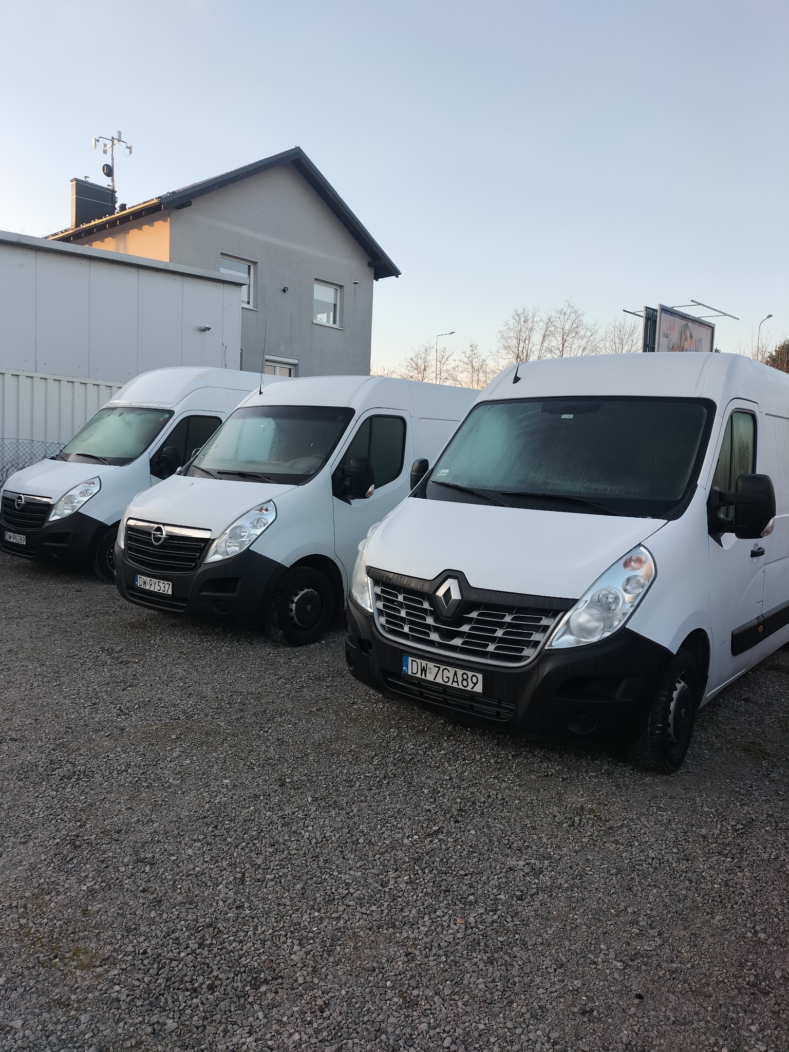 Trzy białe busy dostawcze zaparkowane na żwirowym parkingu przed budynkiem, widoczne tablice rejestracyjne.