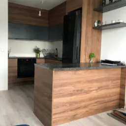 Nowoczesna kuchnia z drewnianymi frontami, grafitowymi szafkami wiszącymi i wyspą z blatem z ciemnego kamienia. Na półkach dekoracje i butelki wina.