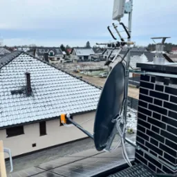Antena satelitarna oraz antena szerokopasmowa zamontowane na dachu domu jednorodzinnego pokrytego panelami fotowoltaicznymi, widok z góry na zaśnieżone dachy sąsiednich domów.