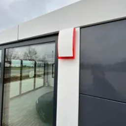 Zewnętrzna sygnalizacja alarmowa z czerwoną lampą błyskową zamontowana na elewacji budynku z szarymi panelami i dużymi oknami.