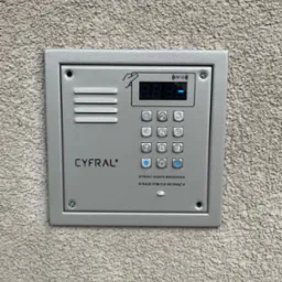 Szary panel domofonu Cyfral z klawiaturą numeryczną i czytnikiem RFID zamontowany na szorstkiej, beżowej ścianie zewnętrznej budynku. Widoczne śruby mocujące panel.