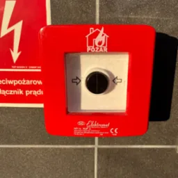 Czerwony, natynkowy przycisk alarmowy pożarowy Elektromet z certyfikatem CNBOP, umieszczony obok tabliczki ostrzegawczej o wysokim napięciu.