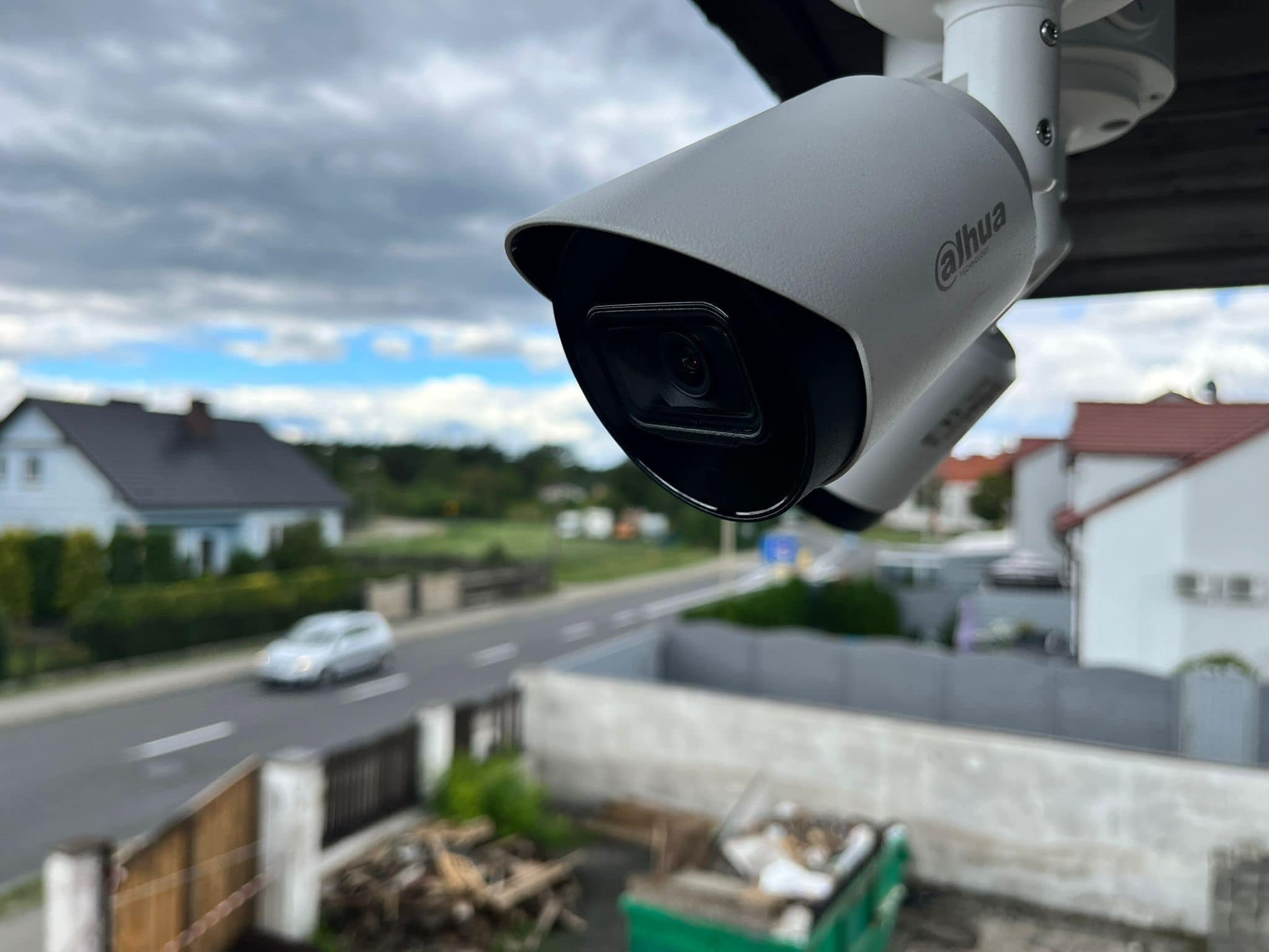 Dwa białe kamery monitoringu Dahua zamontowane pod okapem, widok na ulicę z przejeżdżającym samochodem i domy w tle pod pochmurnym niebem.
