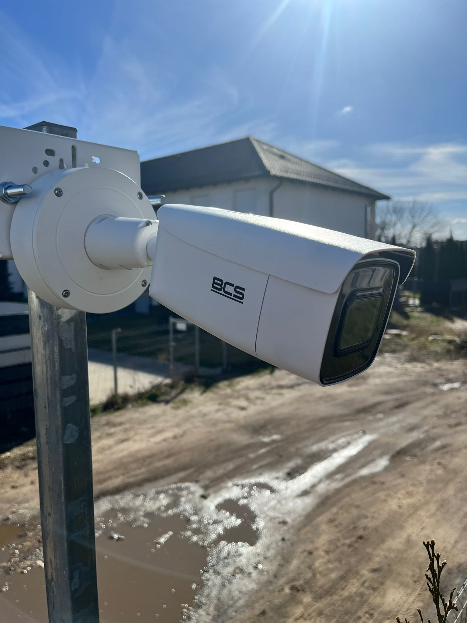 Biała kamera monitoringu BCS zamontowana na metalowym słupku na tle rozlewiska błota i domu jednorodzinnego w słoneczny dzień.