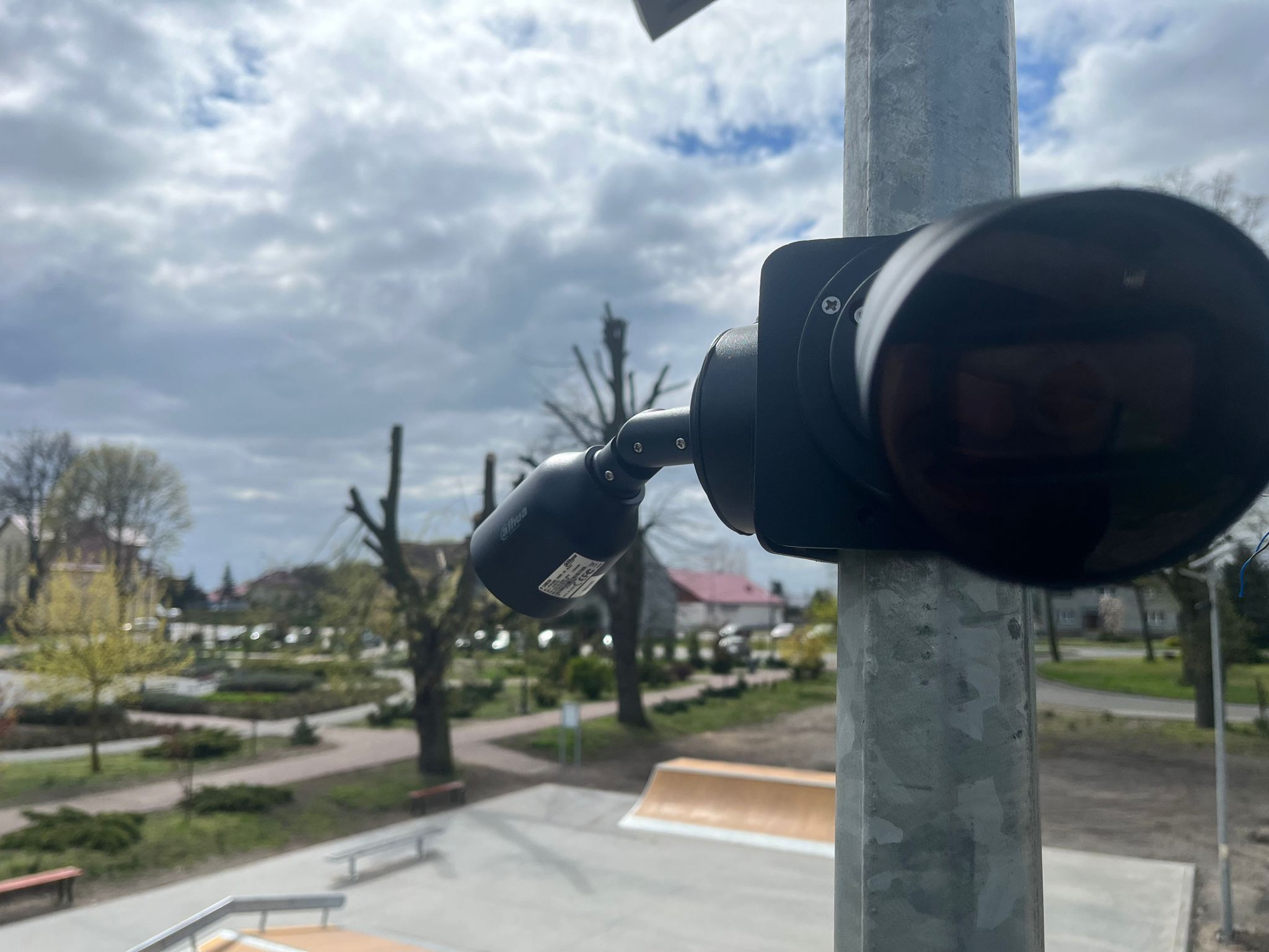 Czarna kamera przemysłowa marki Dahua zamontowana na metalowym słupie, skierowana w stronę skateparku i parku miejskiego w tle, pochmurne niebo.